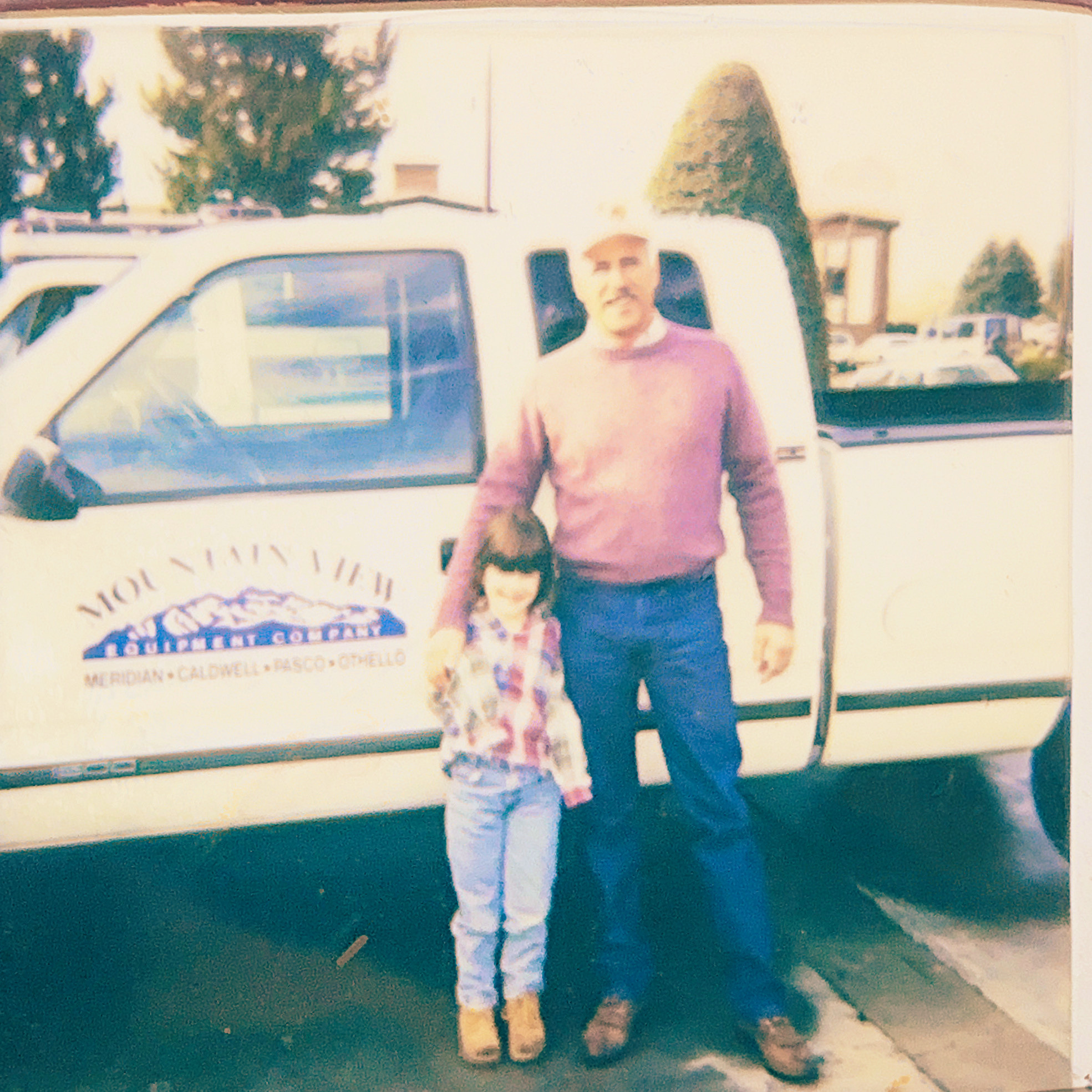A man in a pink sweater and white pants standing outdoors with a young girl in a floral dress. They are near a white vintage vehicle with trees and buildings visible in the background.