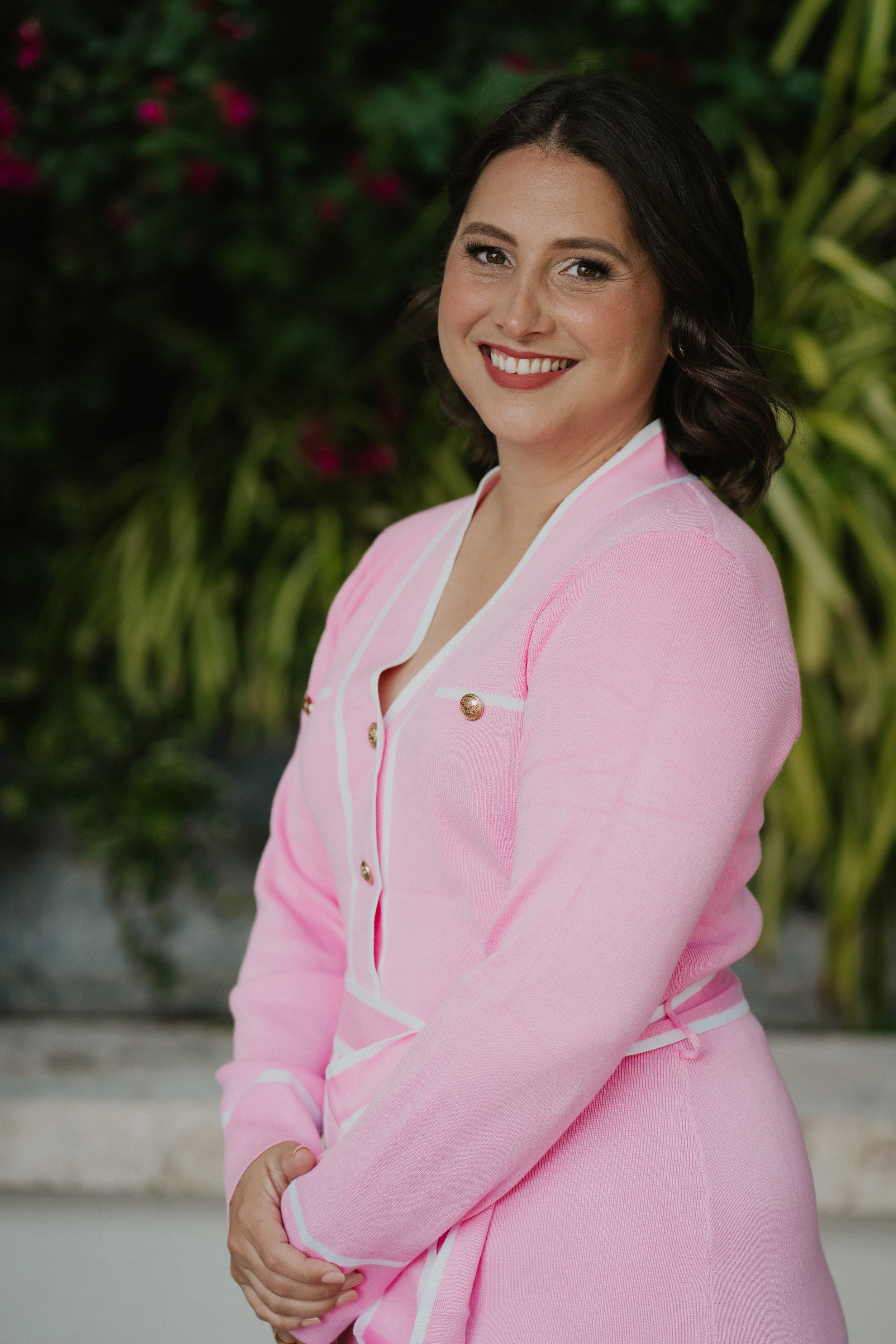 A woman with short dark hair wearing a pink blazer with white trim, smiling and standing outdoors with green foliage in the background.
