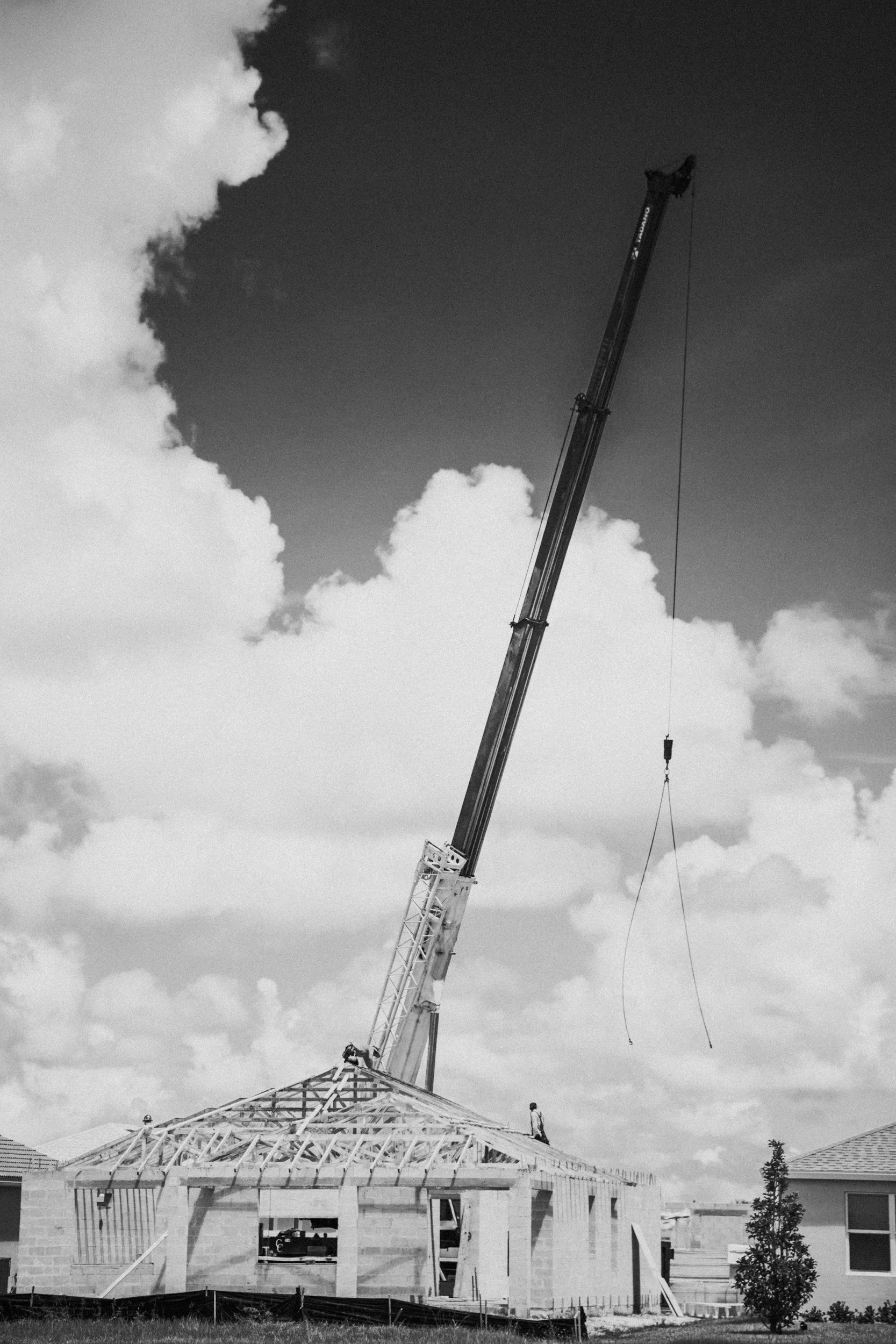 Construction site with a crane lifting a large component, workers on the structure, and a partly built house with a sloped roof, set against a cloudy sky.