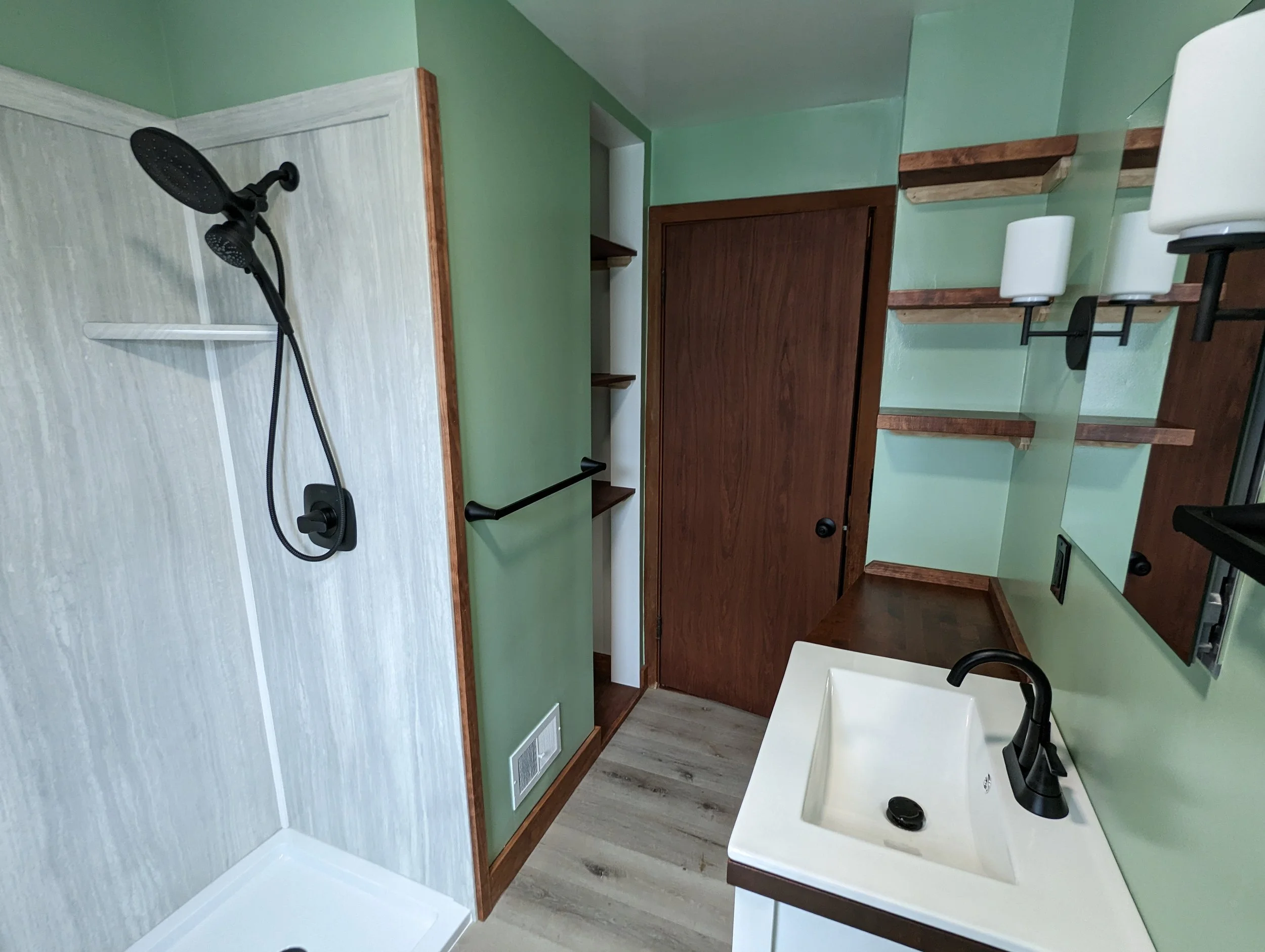 Small bathroom with a walk-in shower, green walls, wooden accents, a white vanity with a black faucet, and a mirror.