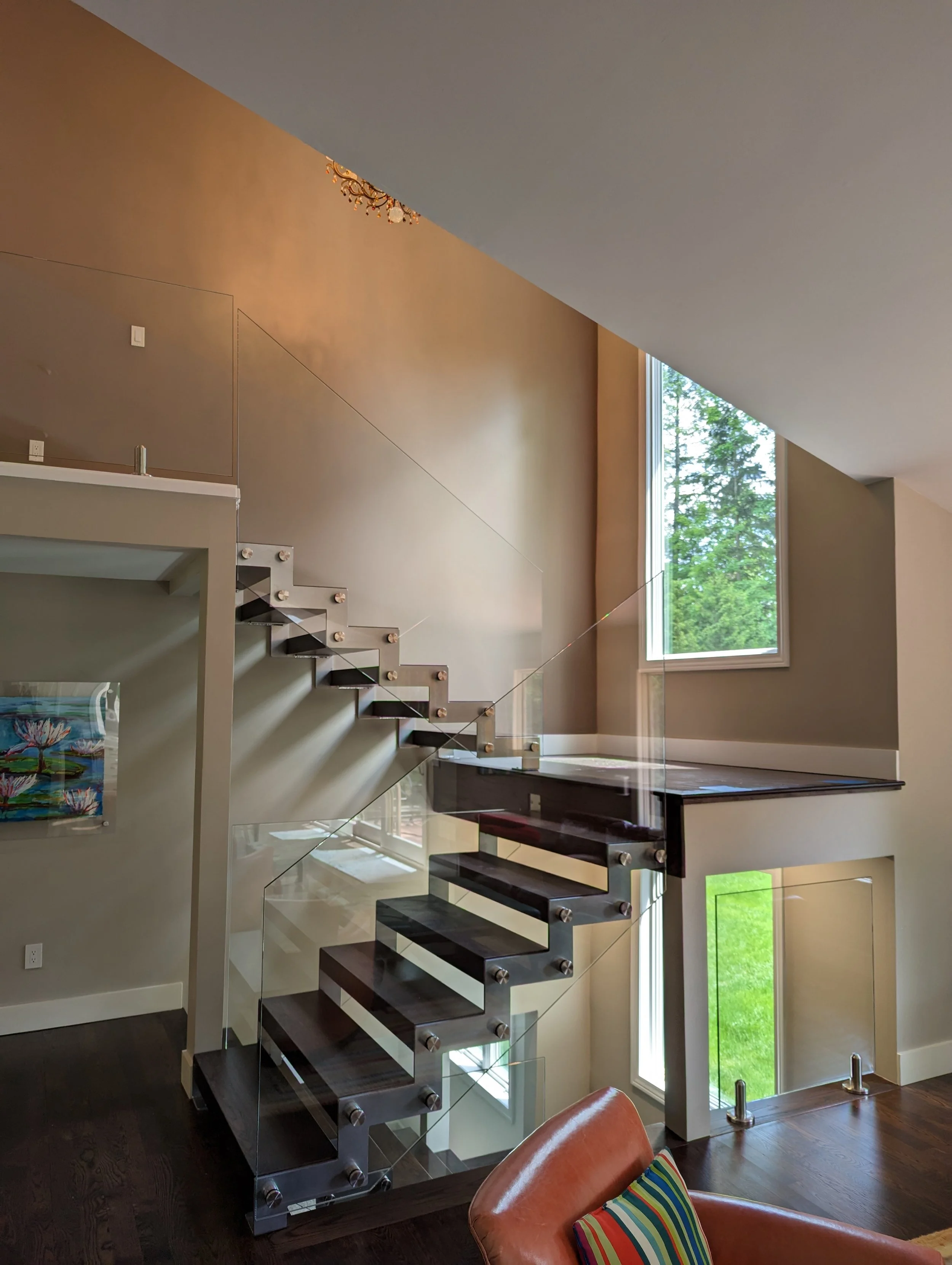 Interior view of a modern home featuring a staircase with dark wood steps and glass railings, large windows with a view of greenery outside, a corner with a colorful painting, and a leather chair with striped cushions.