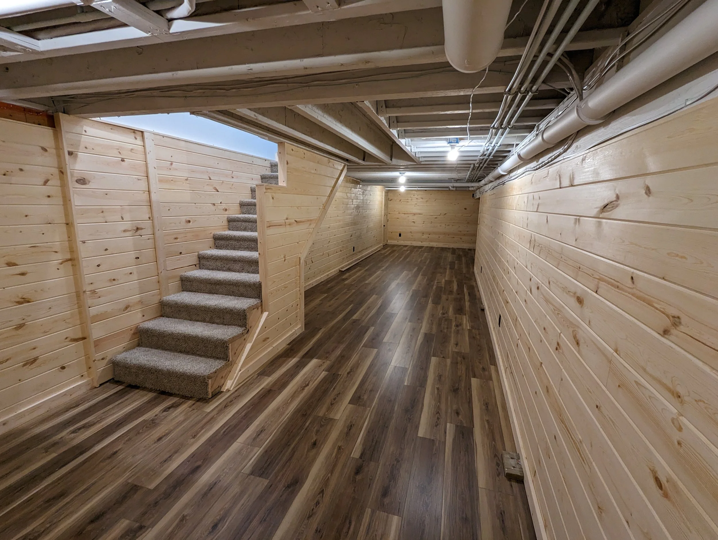 A finished basement with wooden walls, wooden ceiling with exposed beams, and a laminate floor. There are gray carpeted stairs leading up, and several ceiling lights and pipes are visible.