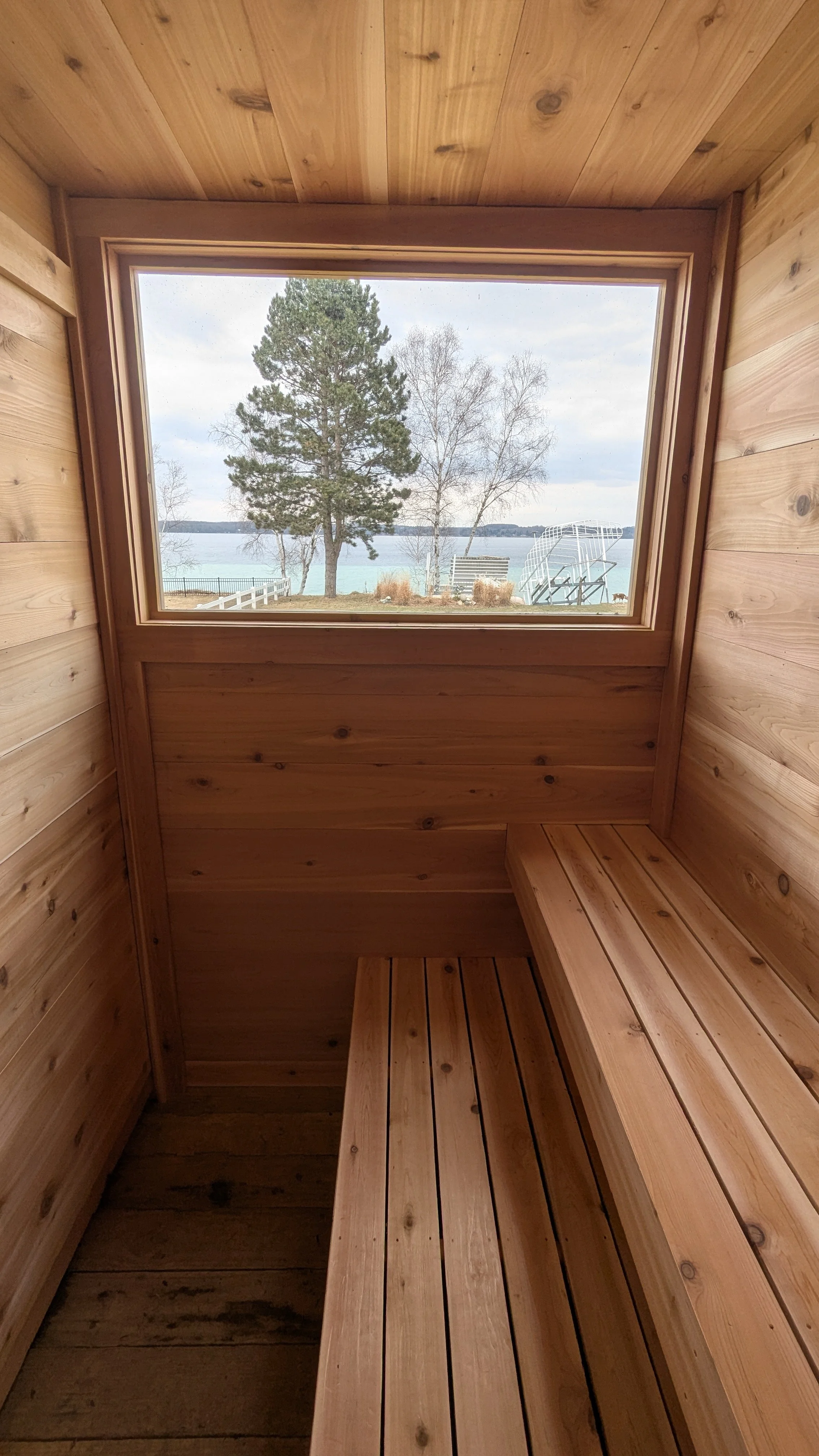 Wooden interior of a small sauna room with a window overlooking trees and water outside.