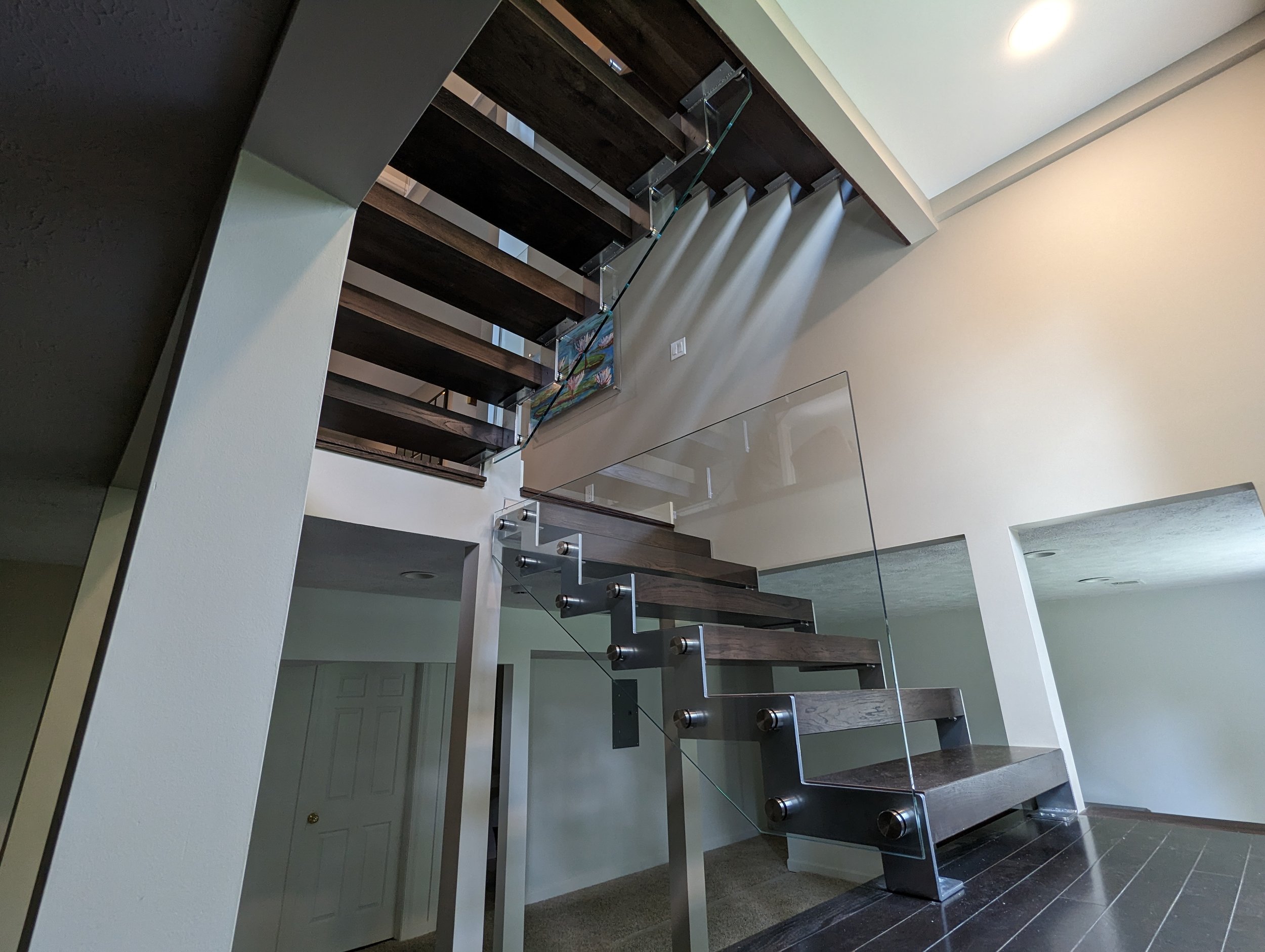Modern staircase with dark wooden steps and glass railings in a spacious home interior, with beige walls and a ceiling light.