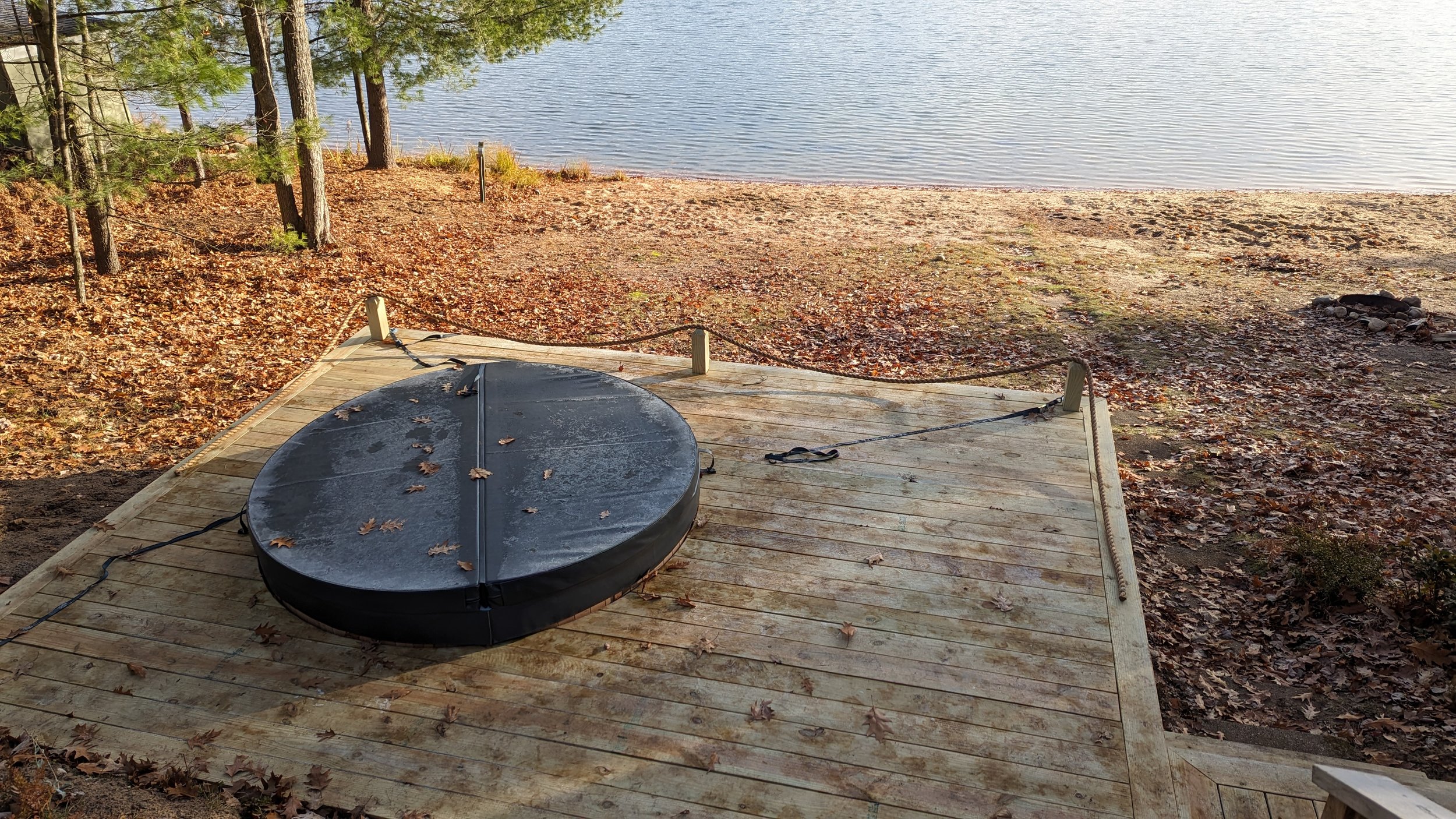 A wooden dock extends into a lake with trees on the shoreline. A covered hot tub is on the dock, covered with fallen leaves. The lake has calm waters, and there is a small fire pit on the sandy beach nearby.