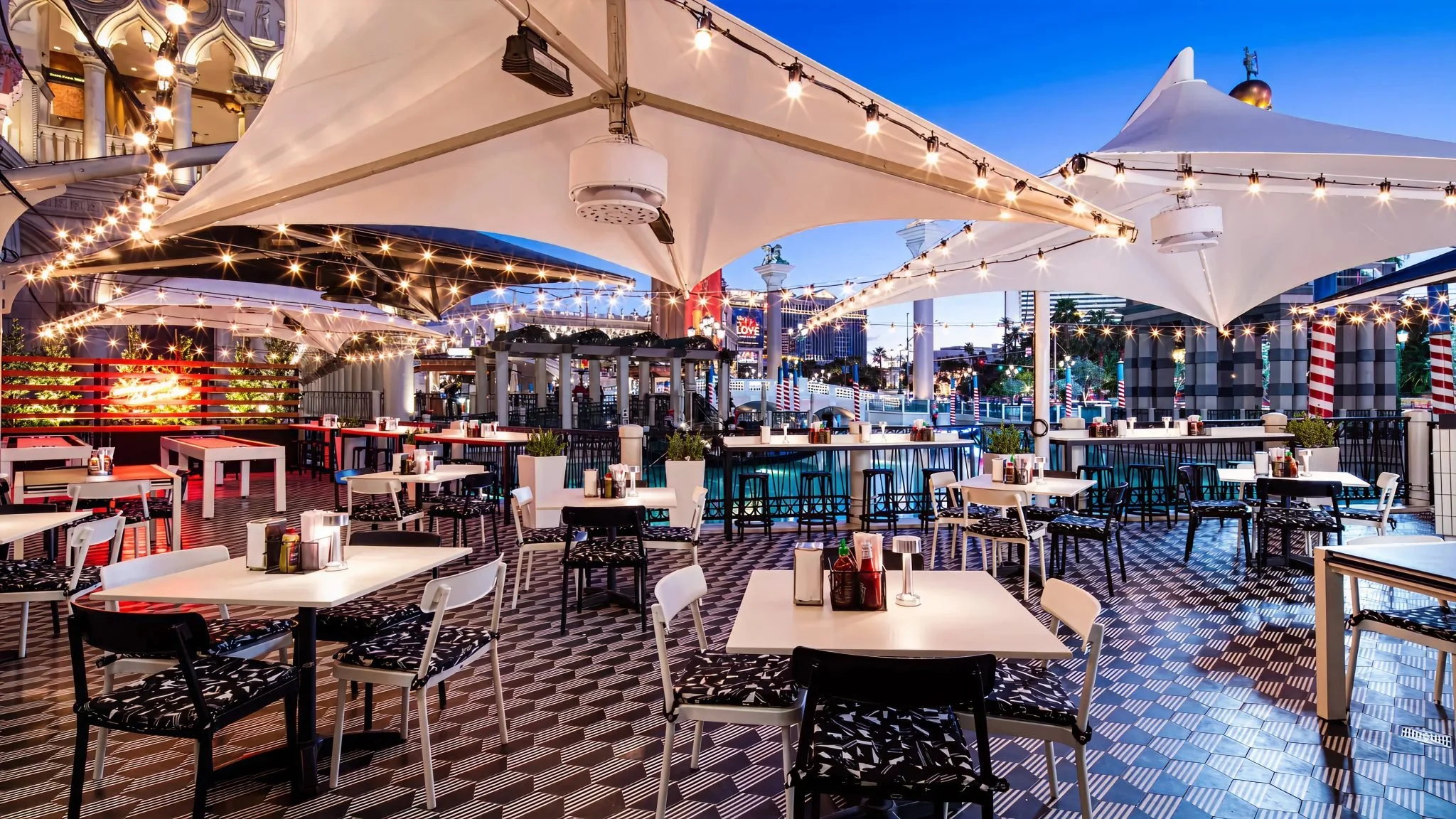 An outdoor dining area with tables and chairs, string lights hanging overhead, large white canopy shades, and a cityscape background with illuminated signs and tall buildings during dusk.