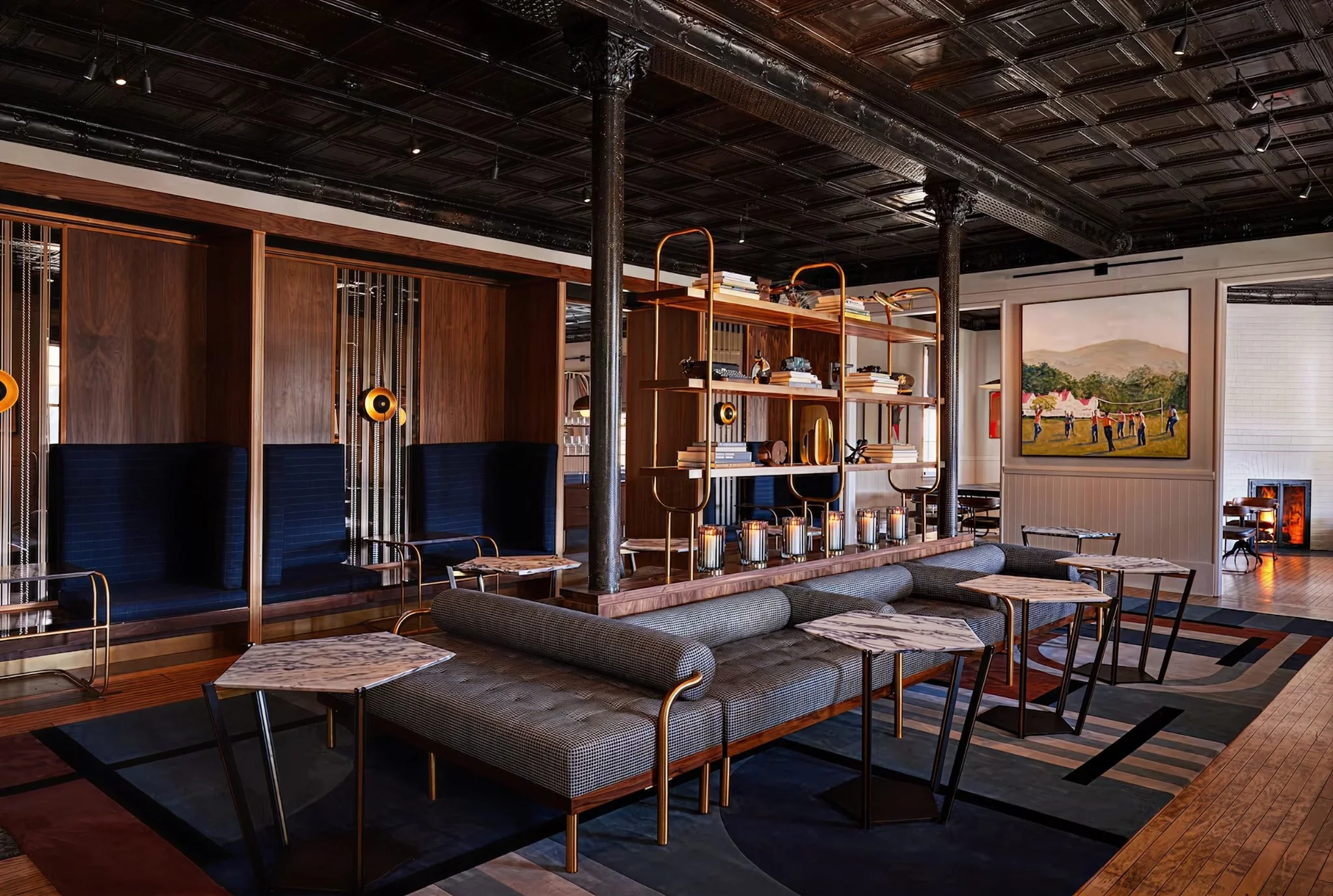 Interior of a stylish room with dark wood ceiling and floors, metal pillars, a wooden shelving unit with books and decor, and a colorful painting of people playing. There are modern chairs, a sitting area with candles, and a large window showing an outdoor scene.