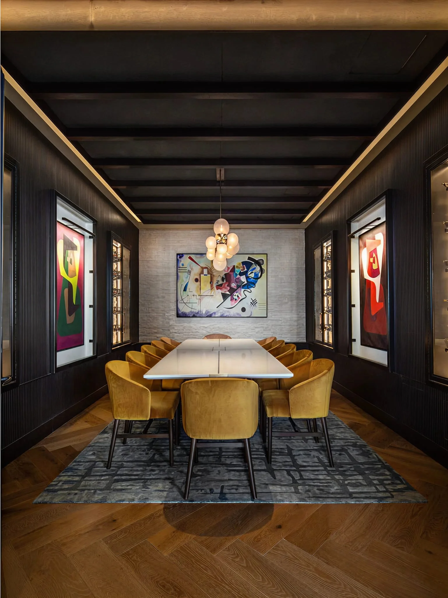 A modern dining room with a long white table, surrounded by yellow upholstered chairs, dark paneling with colorful abstract art on the walls, a rug beneath the table, and a light fixture hanging from the ceiling.