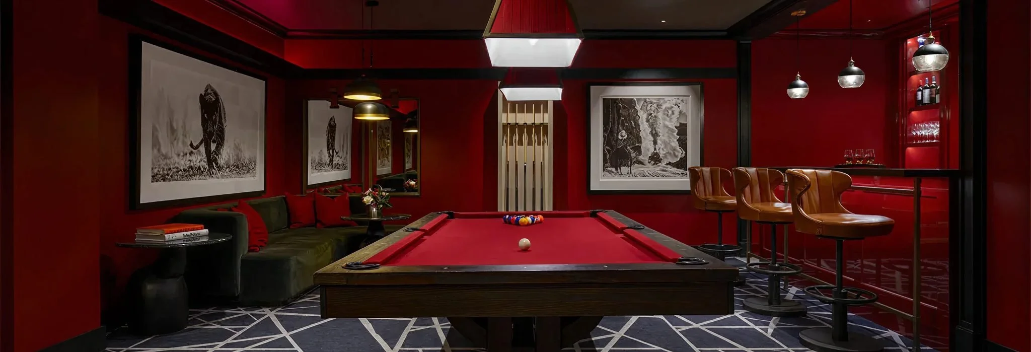 A cozy bar room with red walls, black and white artwork, a pool table with a set of balls and cue ball, a black sofa with red and green pillows, bar stools with brown leather seats, a shelf with bottles and glasses, and decorative lighting.