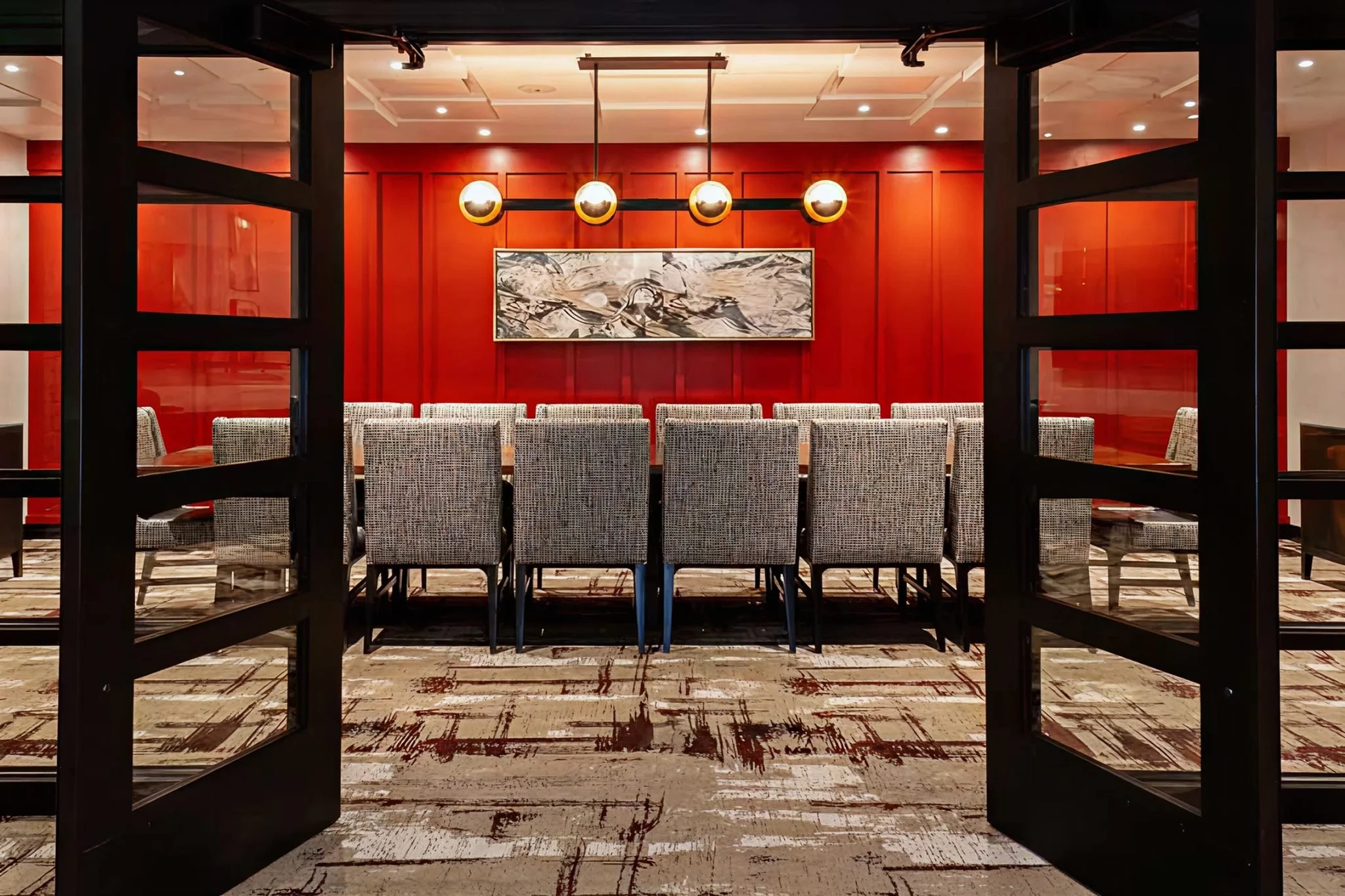 Inside a restaurant or hotel dining room with red walls, a framed painting, patterned chairs, and a modern light fixture hanging from the ceiling. The image is taken through a window or door frame.