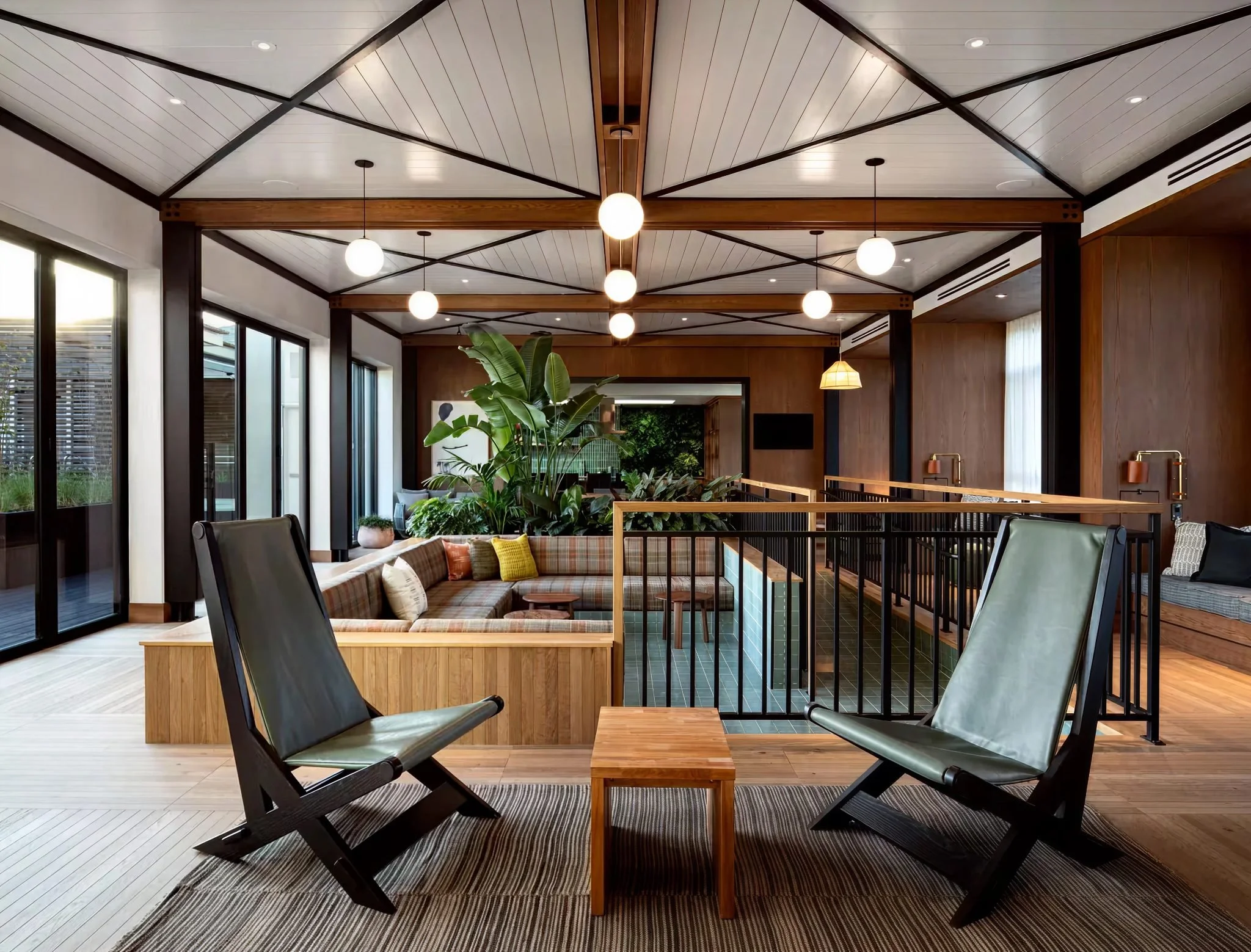 Modern lounge area with black chairs, wooden tables, and green plants under a geometric ceiling with round pendant lights.