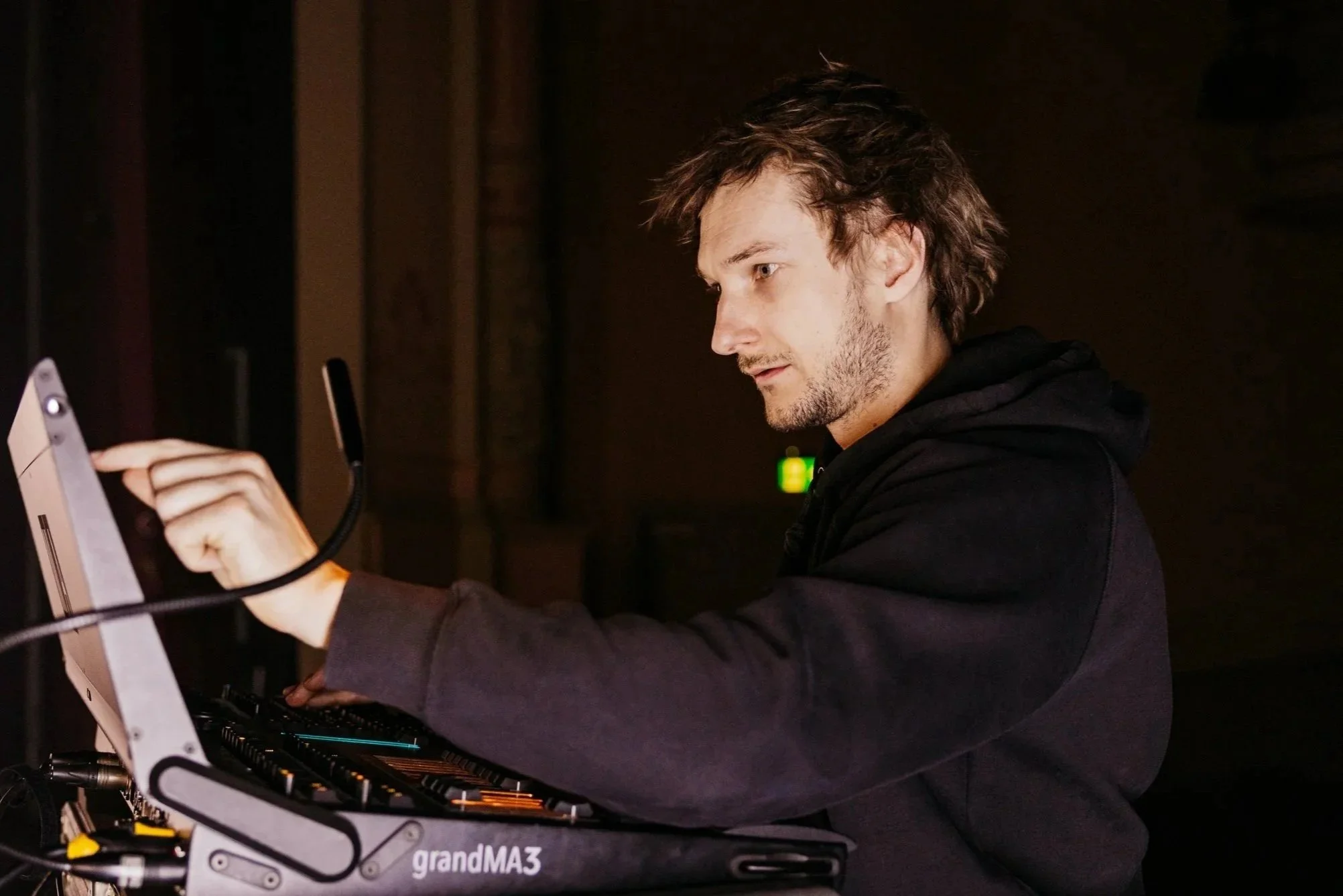 Man met verwarde haar en een lichte baard werkt met een laptop en andere apparatuur, mogelijk in een technologische of creatieve setting.
