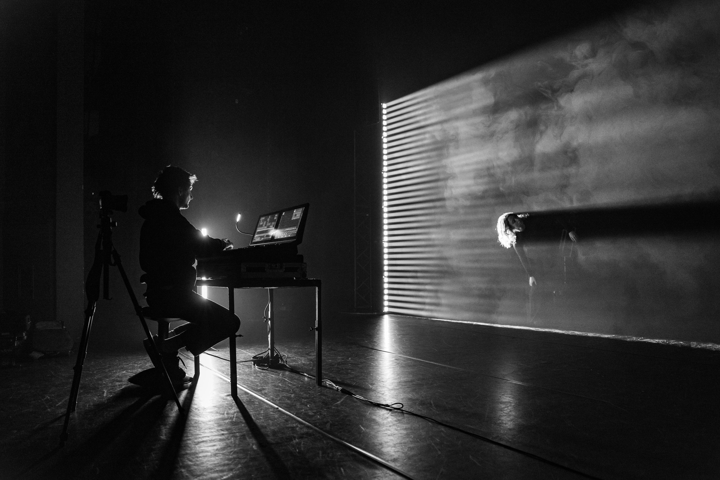 Foto gemaakt door Nathan Reinds. Een persoon die achter een mixconsole zit, naast een andere persoon die op het podium staat, met lichteffecten en rook op de achtergrond.