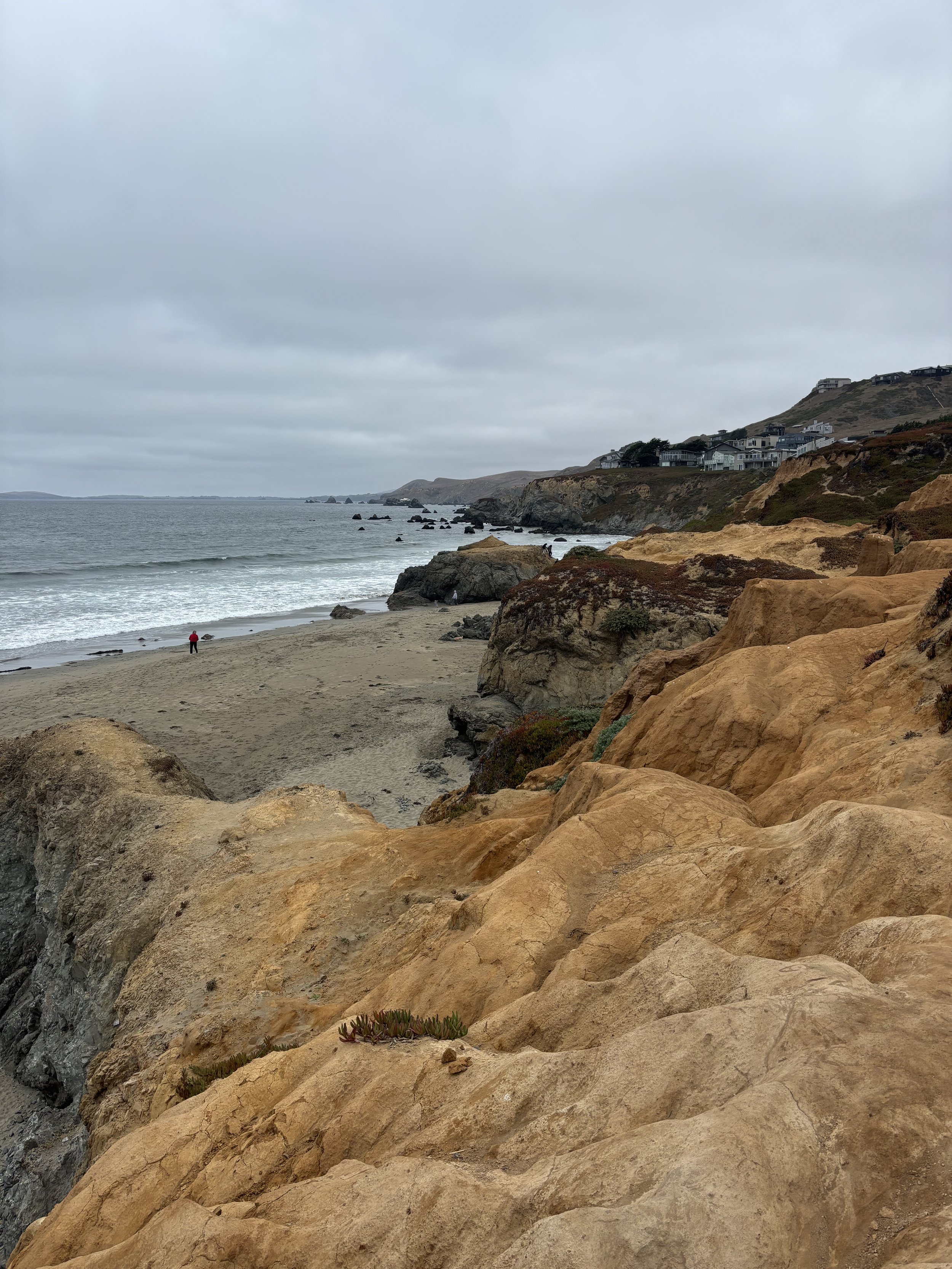 Overcast beach scene with large sandy and rocky cliffs in the foreground, ocean waves, and a small figure walking on the beach, with hillside houses in the background.