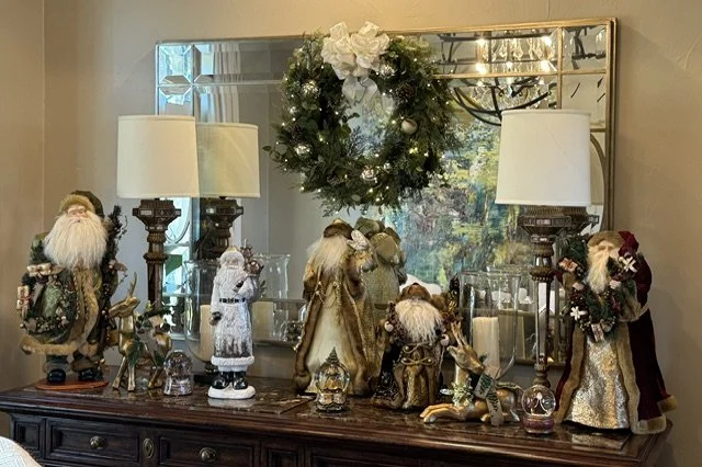 A decorated Christmas display on a dark wooden sideboard, featuring Santa and angel figurines, glass ornaments, candles, and a Christmas wreath with white flowers hanging in the mirror's reflection, flanked by two table lamps.