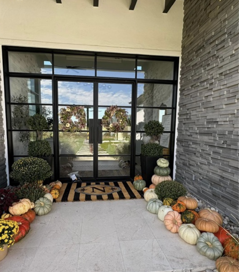 Entrance with glass door decorated with floral wreaths, surrounded by pumpkins and potted plants, with a decorative doormat in front.