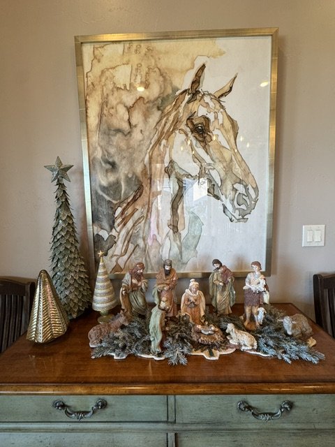 Decorative nativity scene with figurines of people and animals on a wooden surface, in front of a large framed horse watercolor painting, with Christmas trees on the left side.