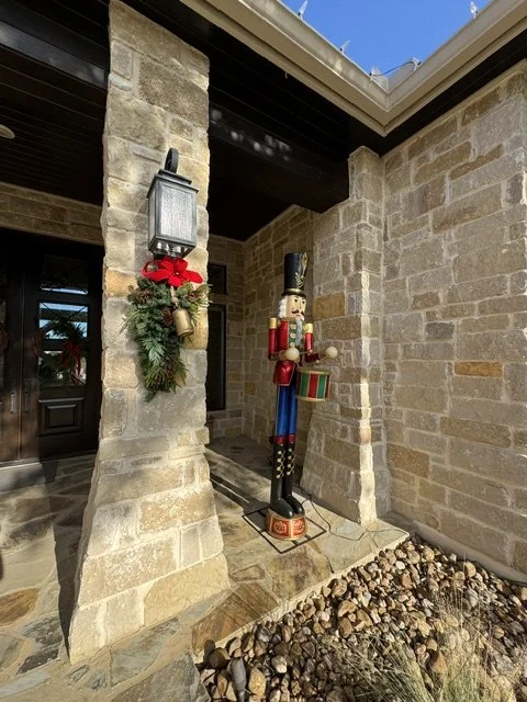 Decorative plastic toy soldier with drum standing outside house, next to stone wall with wall-mounted lantern decorated with holiday wreath and red bow, on a porch with rocky gravel ground.