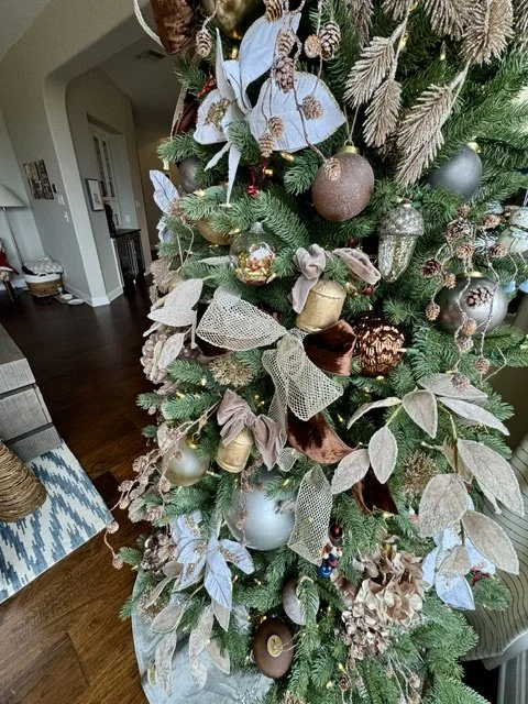 Close-up of a decorated Christmas tree with gold, silver, and brown ornaments, ribbons, and faux foliage, in a living room with hardwood floors.