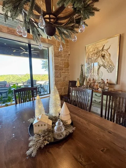 Christmas-themed dining room table with a centerpiece of miniature white Christmas trees, cones, and garland. A chandelier decorated with greenery and glass ornaments hangs above. A large window shows an outdoor patio and greenery. In the background,