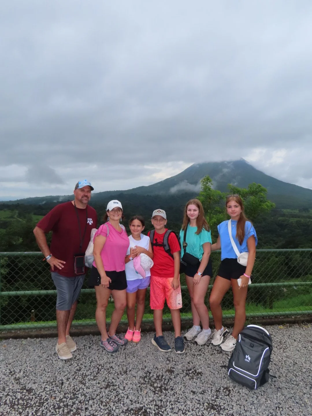 Group of six people, including two adults and four children, standing on a gravel area with a mountain and forest in the background during cloudy weather.