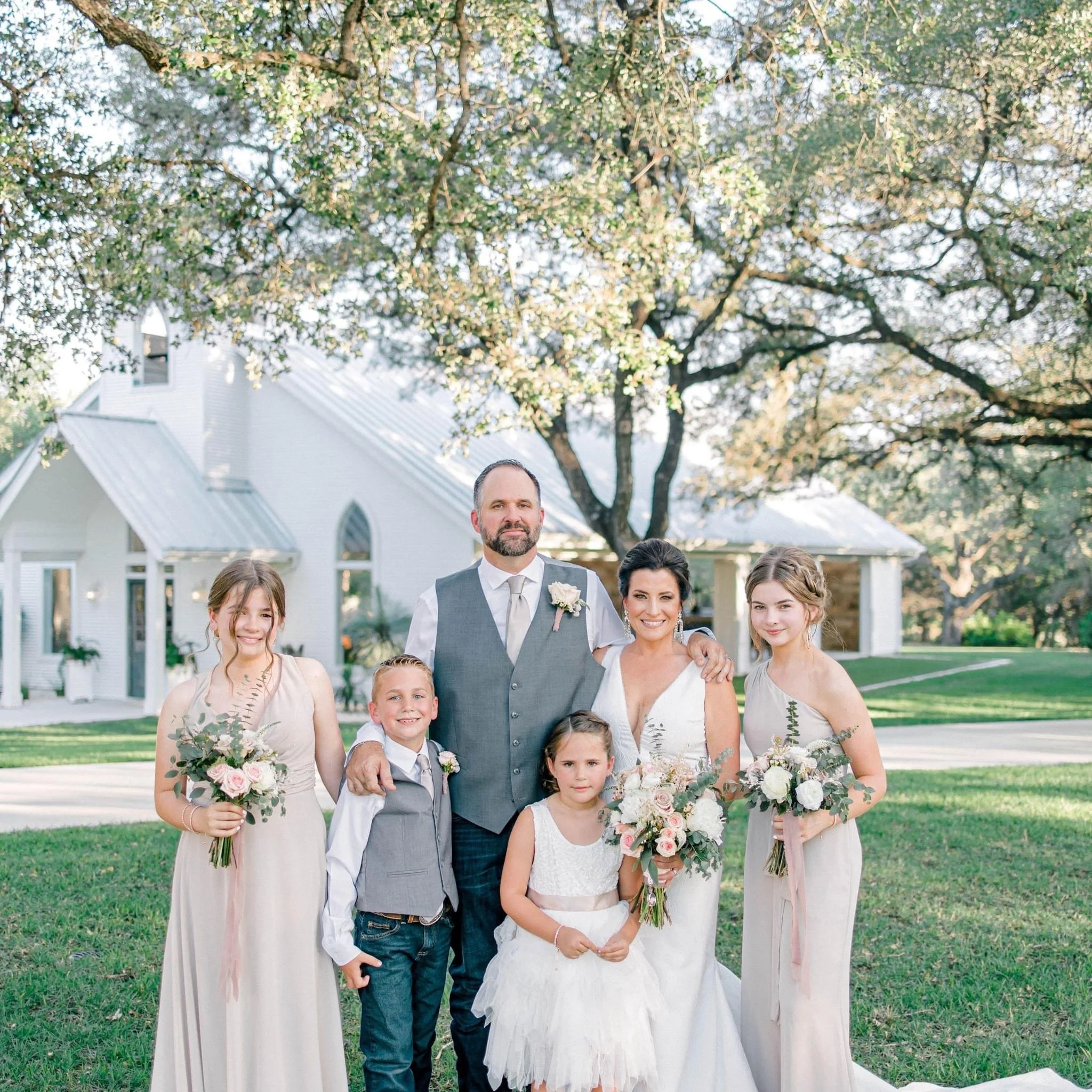 Group of people at a wedding outside, including children and adults, with trees and a white house in the background.
