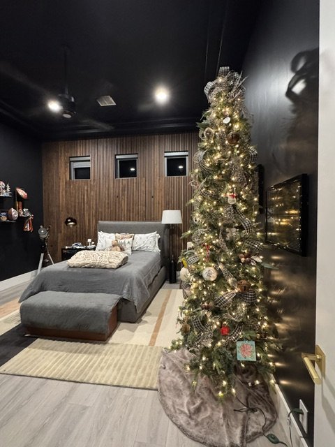 A bedroom decorated for Christmas with a Christmas tree illuminated with lights and ornaments next to a wall, with a bed, pillows, a lamp, and wall decorations in the background.