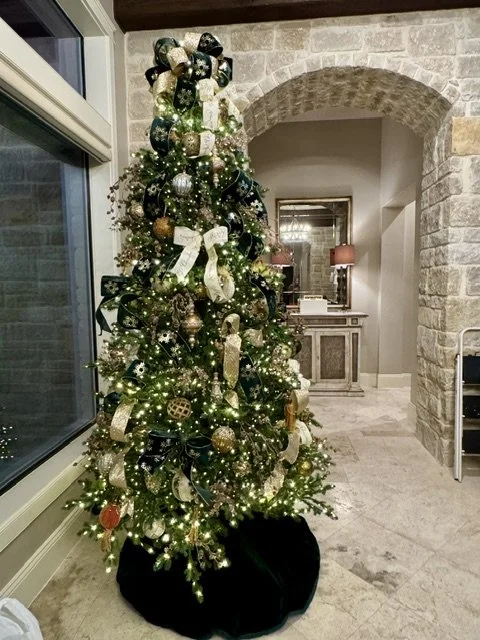 Decorated Christmas tree with gold, white, and green ornaments, ribbons, and lights in a decorated indoor space.
