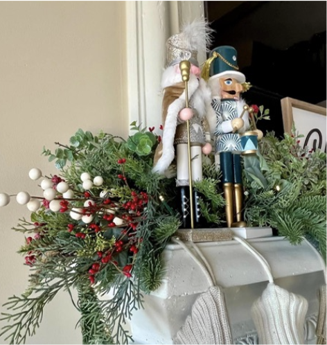 Festive holiday arrangement with greenery, red berries, and white berries, topped with two decorative nutcracker figures and a toy soldier, placed on a mantelpiece with Christmas stockings.