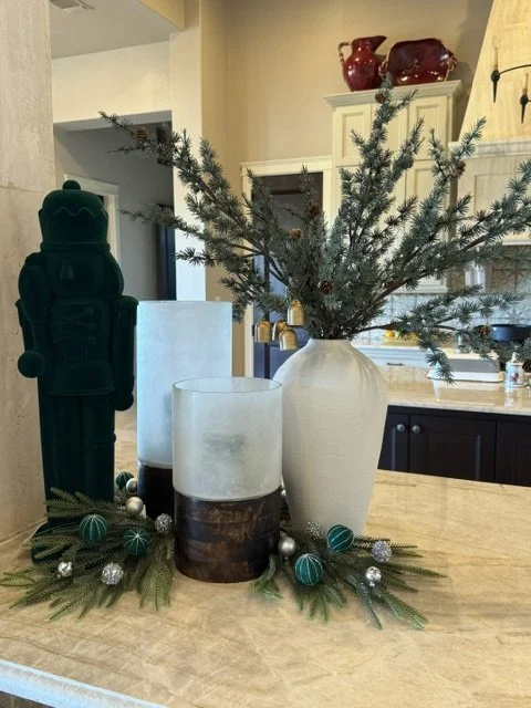 Holiday centerpiece with pine branches in a white vase, candles, and decorative ornaments on a kitchen counter.