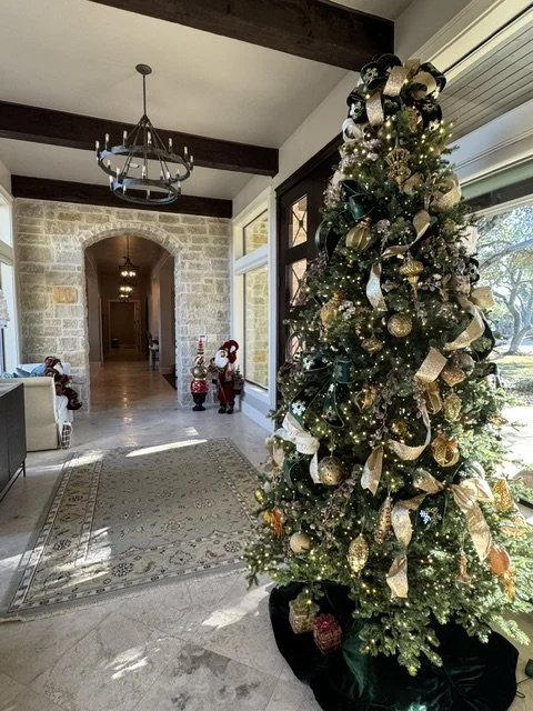 Decorated Christmas tree with gold and green ribbons, ornaments, and lights in a bright living room with large windows, a chandelier, and holiday decorations.
