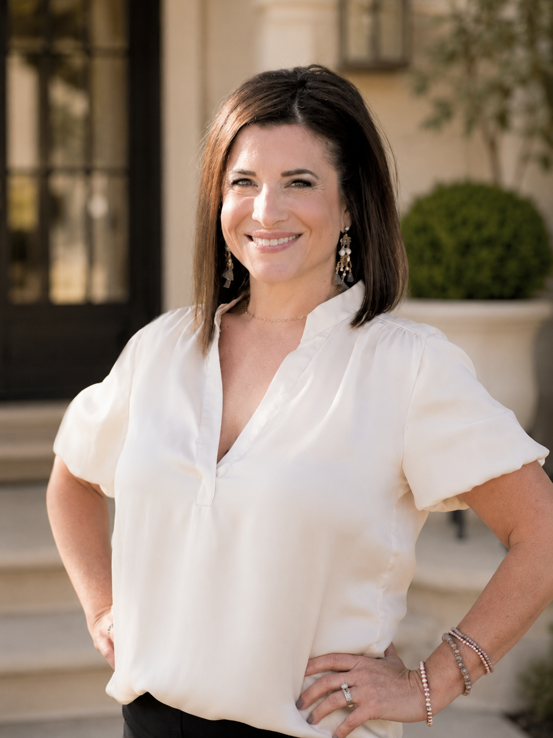 A woman with shoulder-length dark brown hair, smiling and standing outdoors in front of a building with steps and greenery, wearing a white blouse and jewelry.
