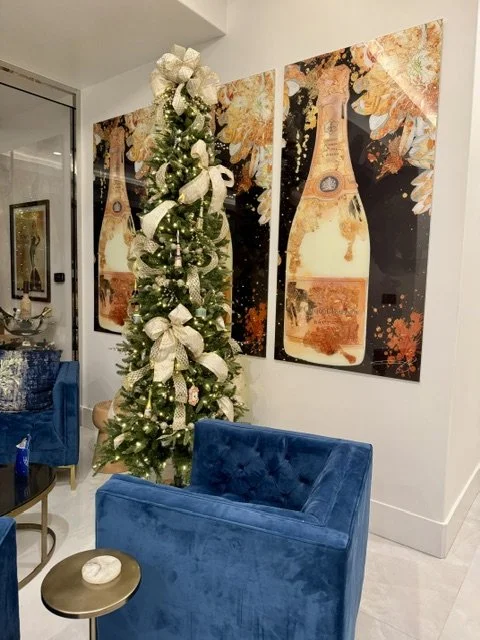 Decorated holiday Christmas tree with white ribbons and ornaments in an indoor living space, with blue velvet chairs and festive artwork on the wall.