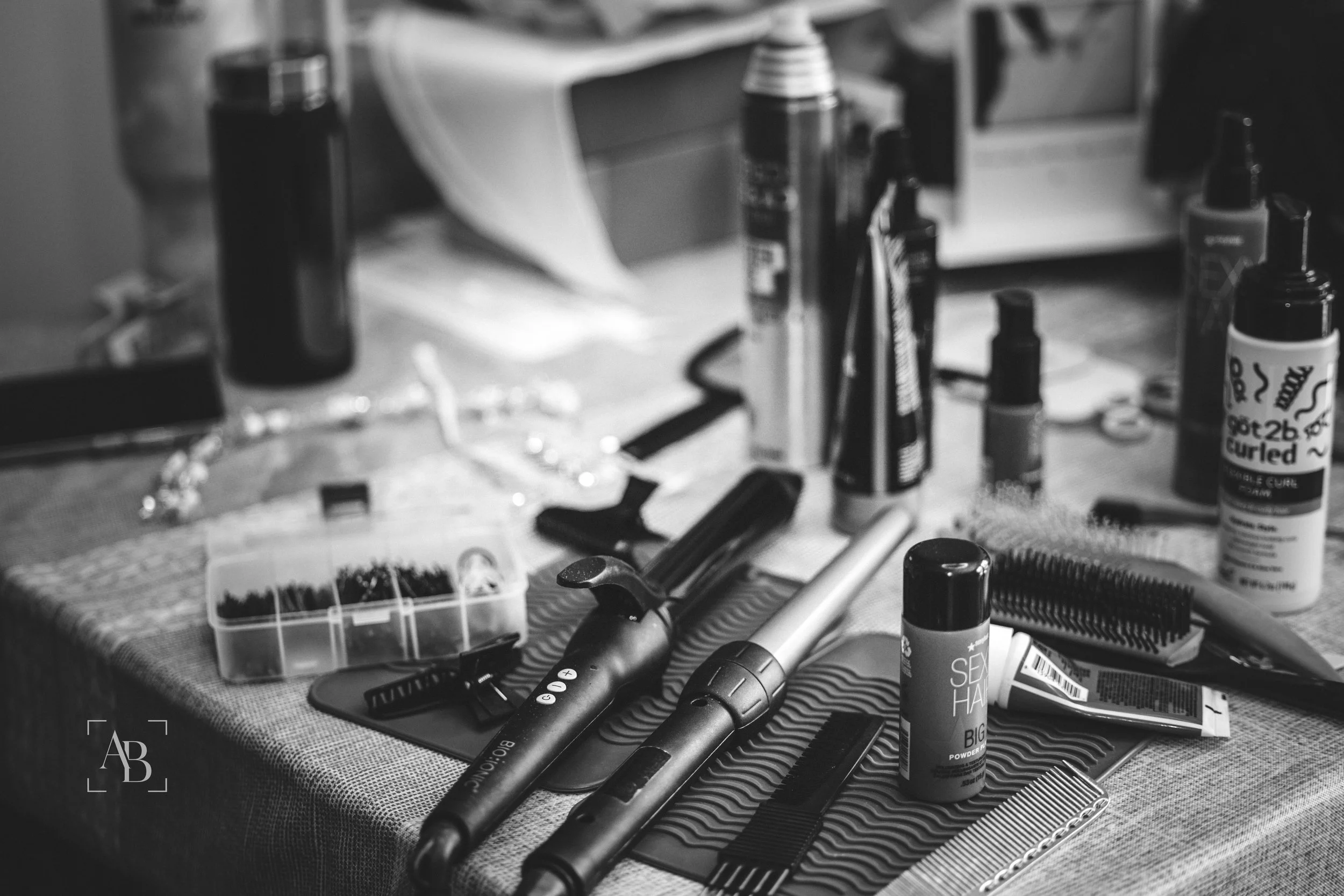 A cluttered table with various hair styling tools and products, including spray bottles, a hair comb, a flat iron, trimmers, and bottles of hair spray, surrounded by scattered small items.