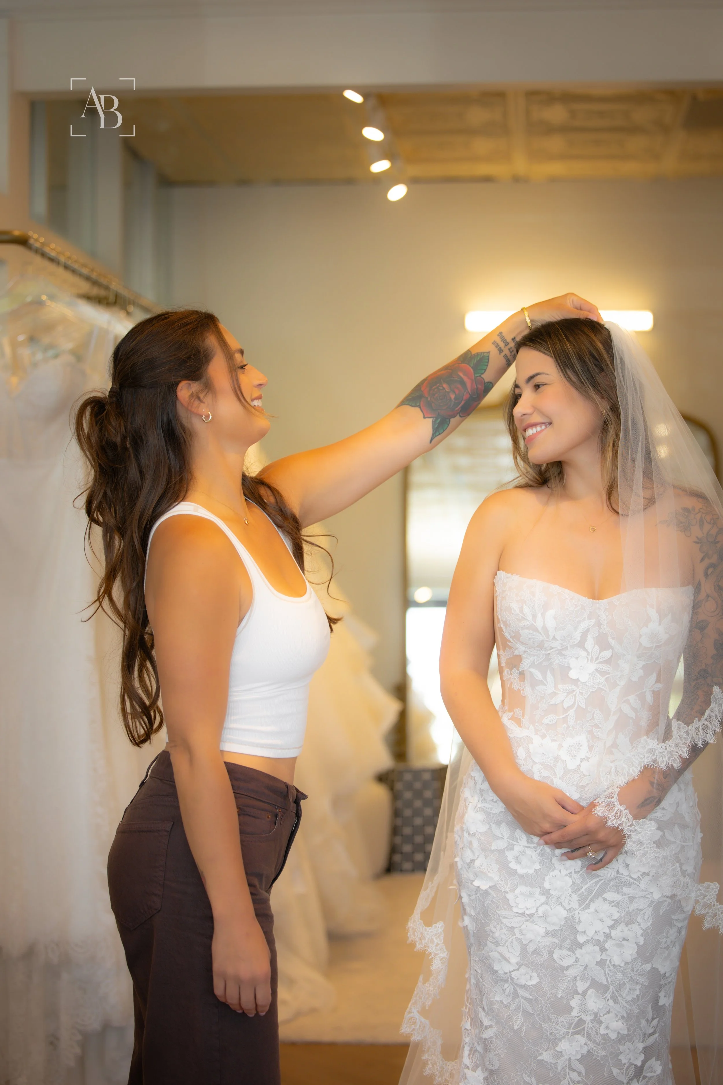 Two women on their wedding day, one dressed in a white wedding gown with a veil, the other in a white tank top and brown pants. The woman in the wedding gown is smiling while the other is adjusting her veil.
