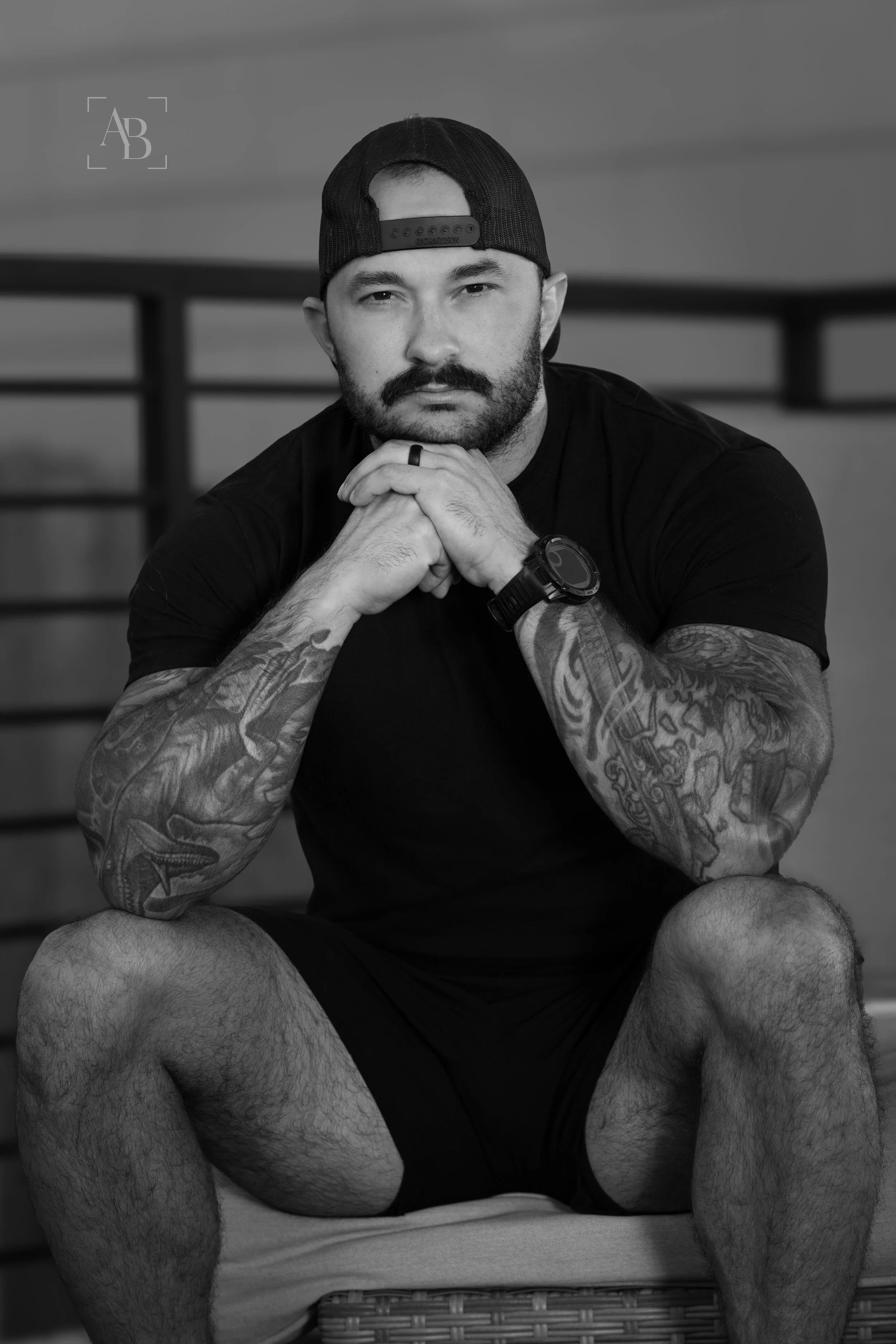 A black and white portrait of a man with a beard and tattoos, wearing a backward baseball cap and casual sportswear, sitting outdoors.