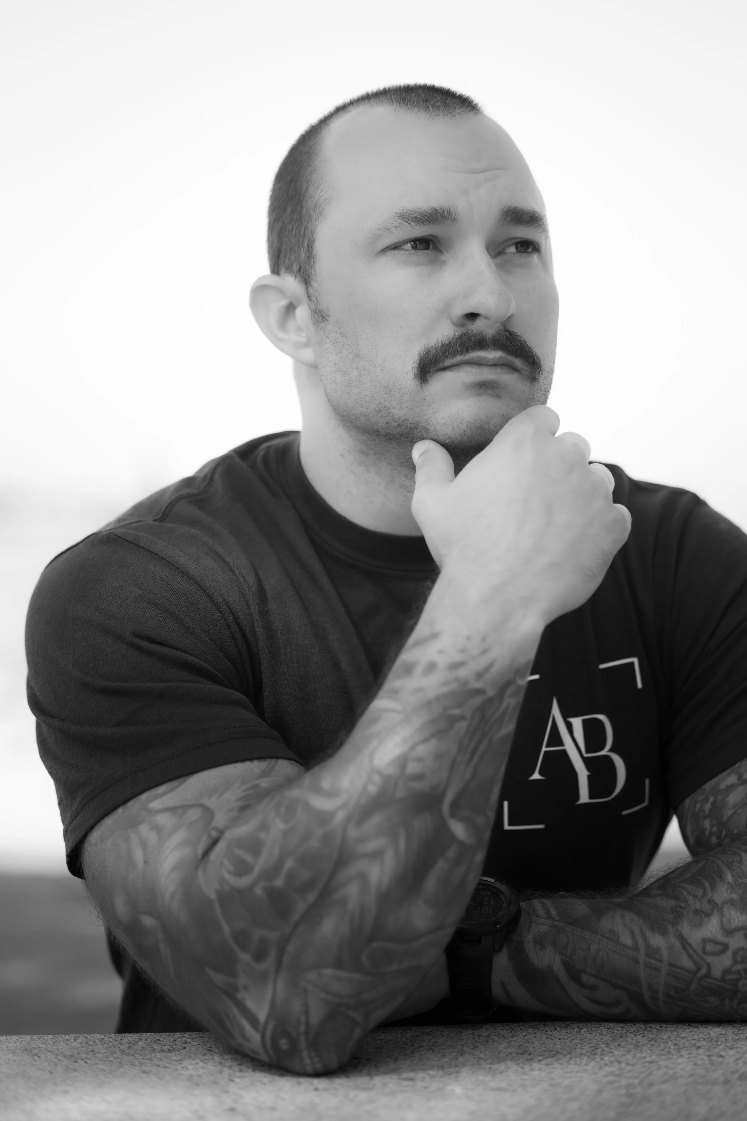 Black and white photo of a man with short hair, a mustache, and tattoos on his arm, resting his chin on his hand, wearing a shirt with the letters 'AB' and a square border.