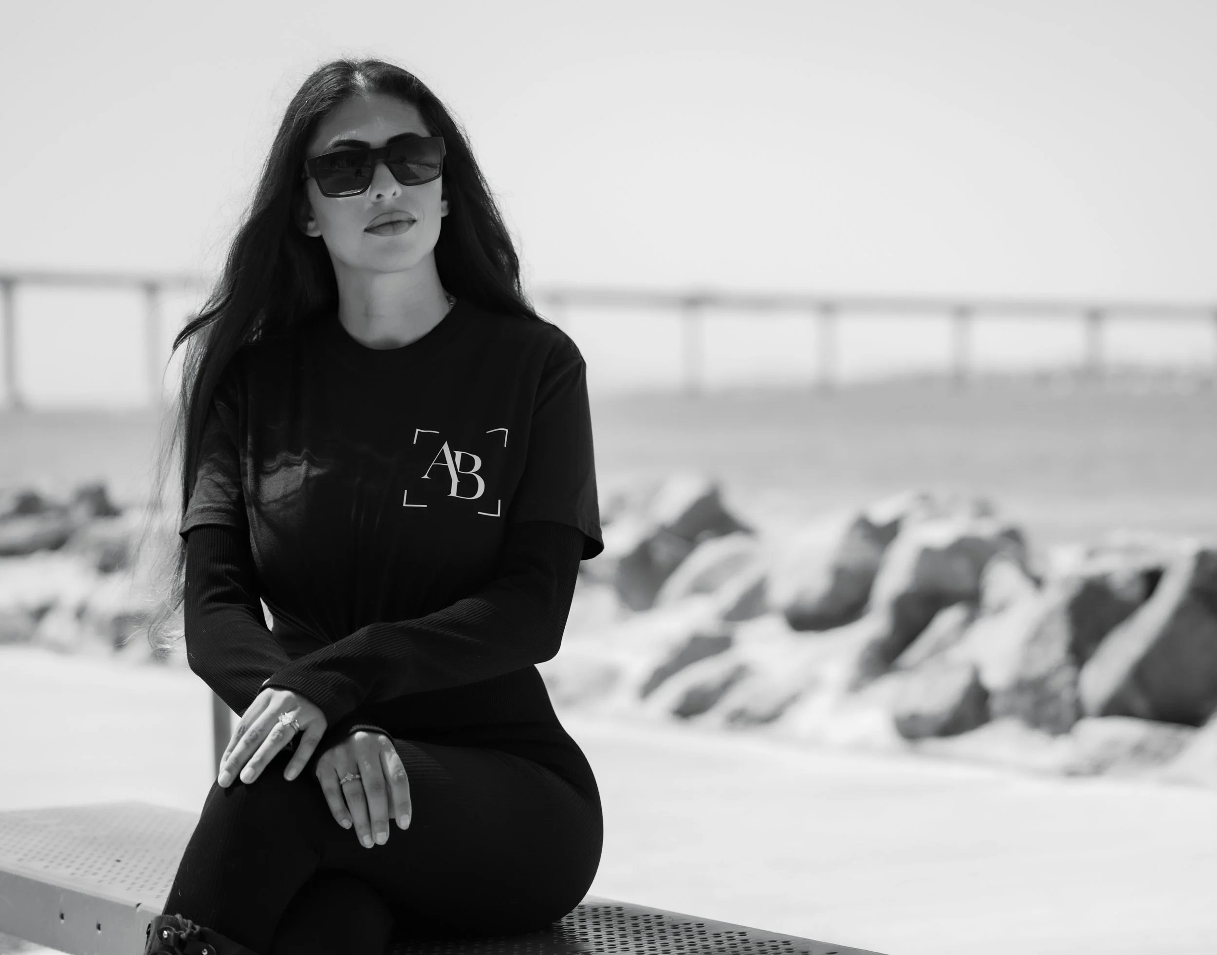 A woman in sunglasses and black clothing sitting on a bench outdoors in a snowy environment.