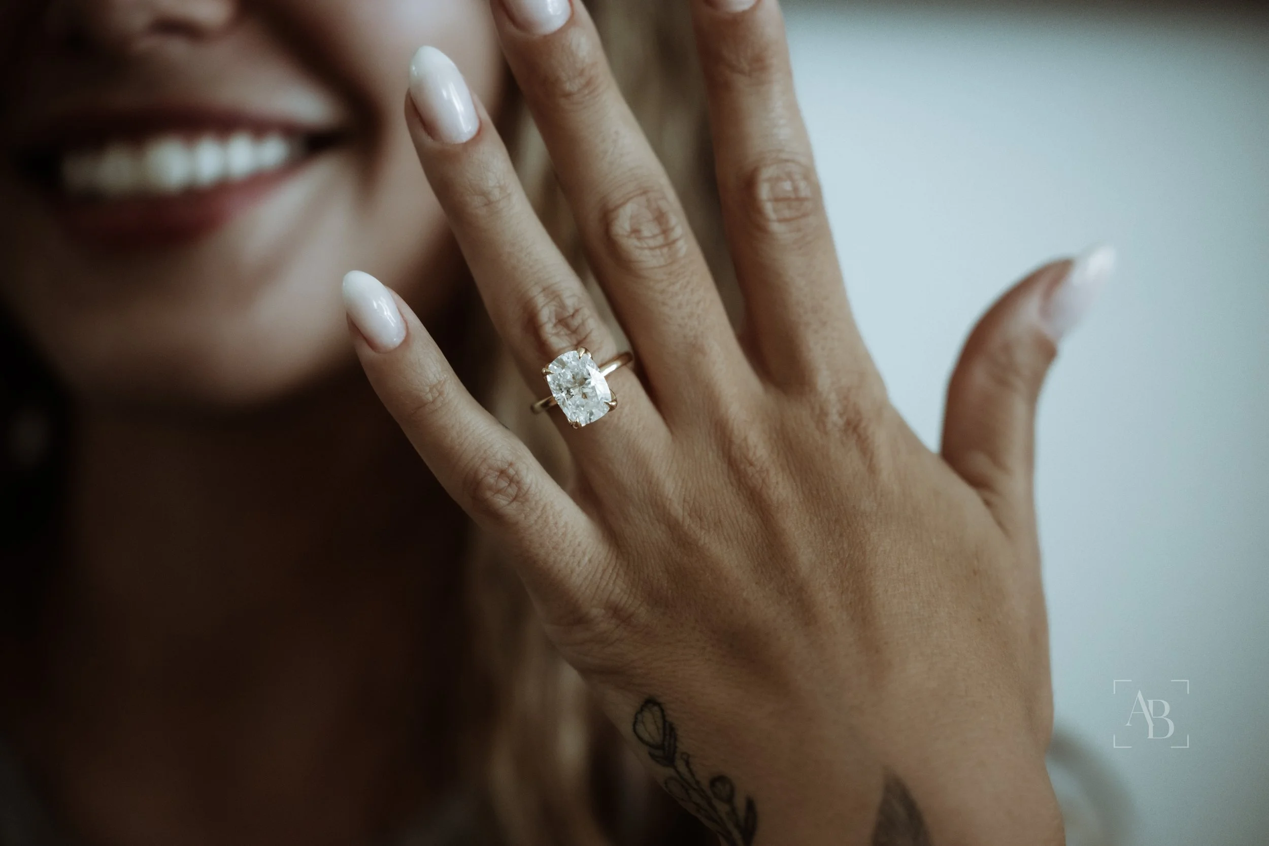 Close-up of a woman's face partially visible, showing her smiling mouth and teeth. Her hand is held up near her face, displaying an elegant diamond ring on her ring finger. The woman has light brown or tan skin and well-manicured nails with a light, natural polish. The background is plain and neutral.