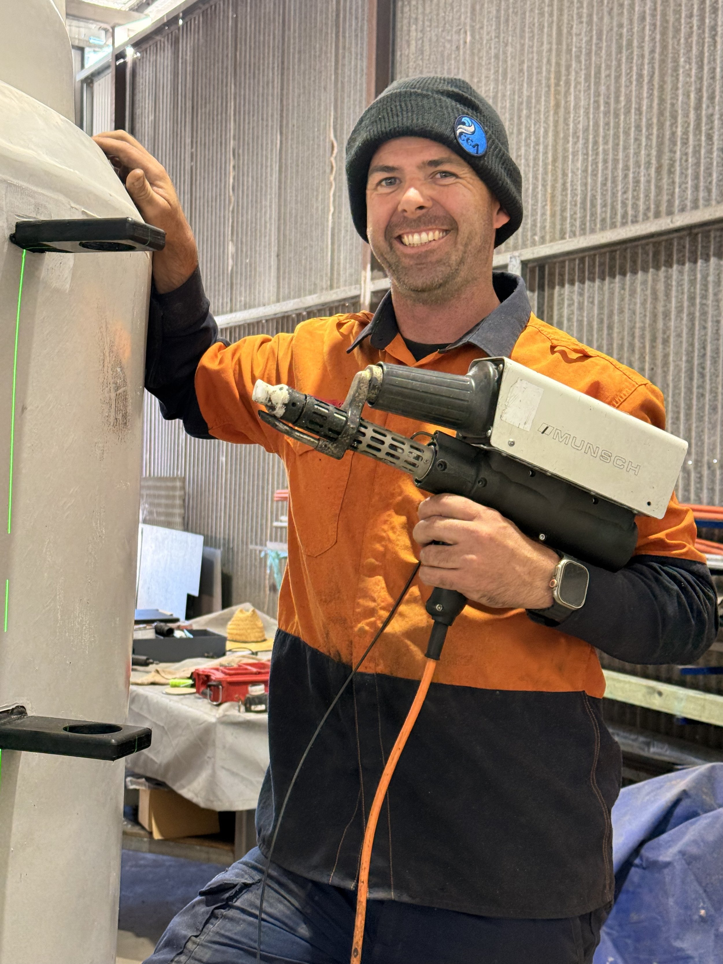 A smiling man wearing a black beanie, orange jacket, and black shirt, working with a MIG welding machine in an industrial workshop.