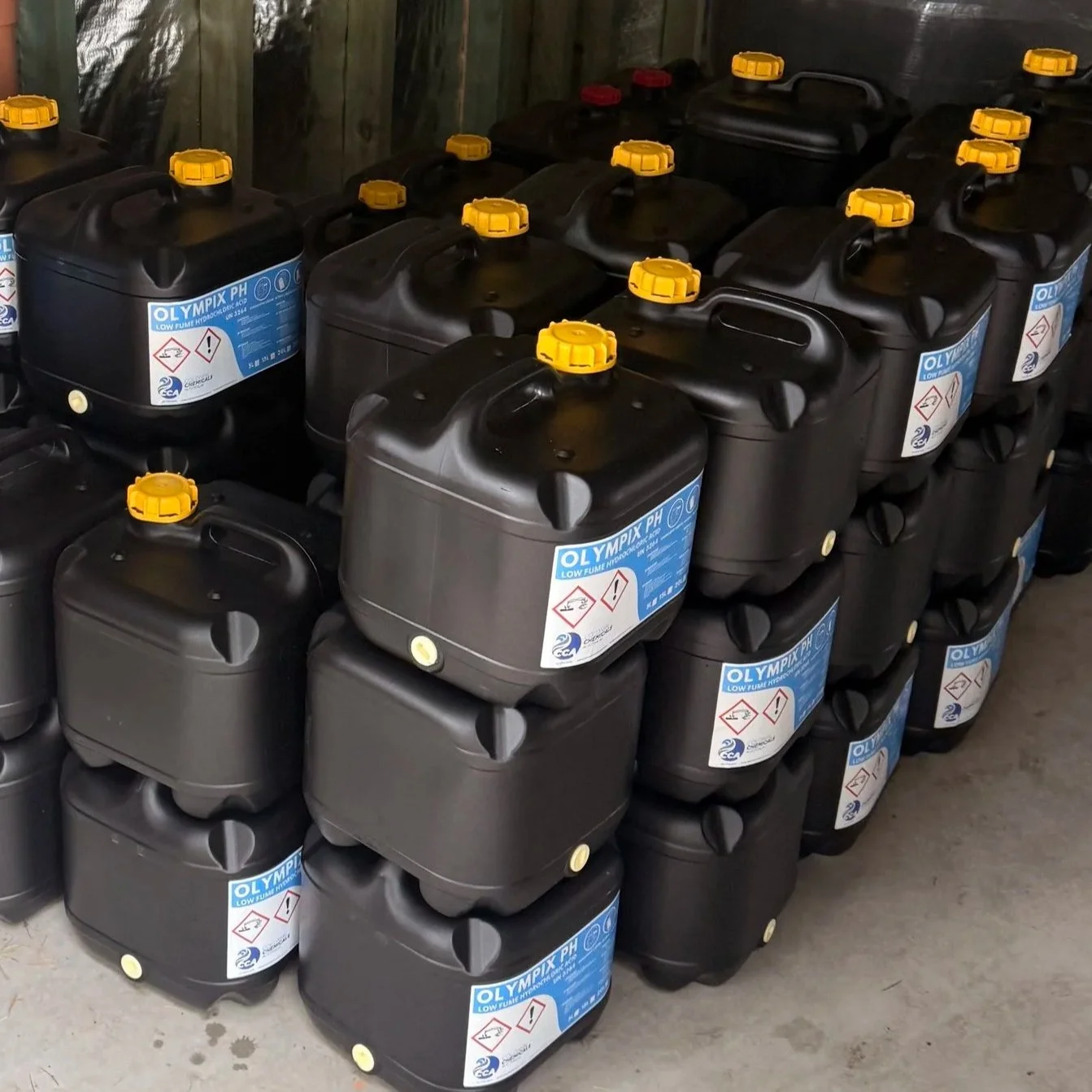 Stacked black plastic containers with yellow caps, labeled 'Olympix PH' for low-fluoride hydrofluoric acid, in a storage area with wooden walls.