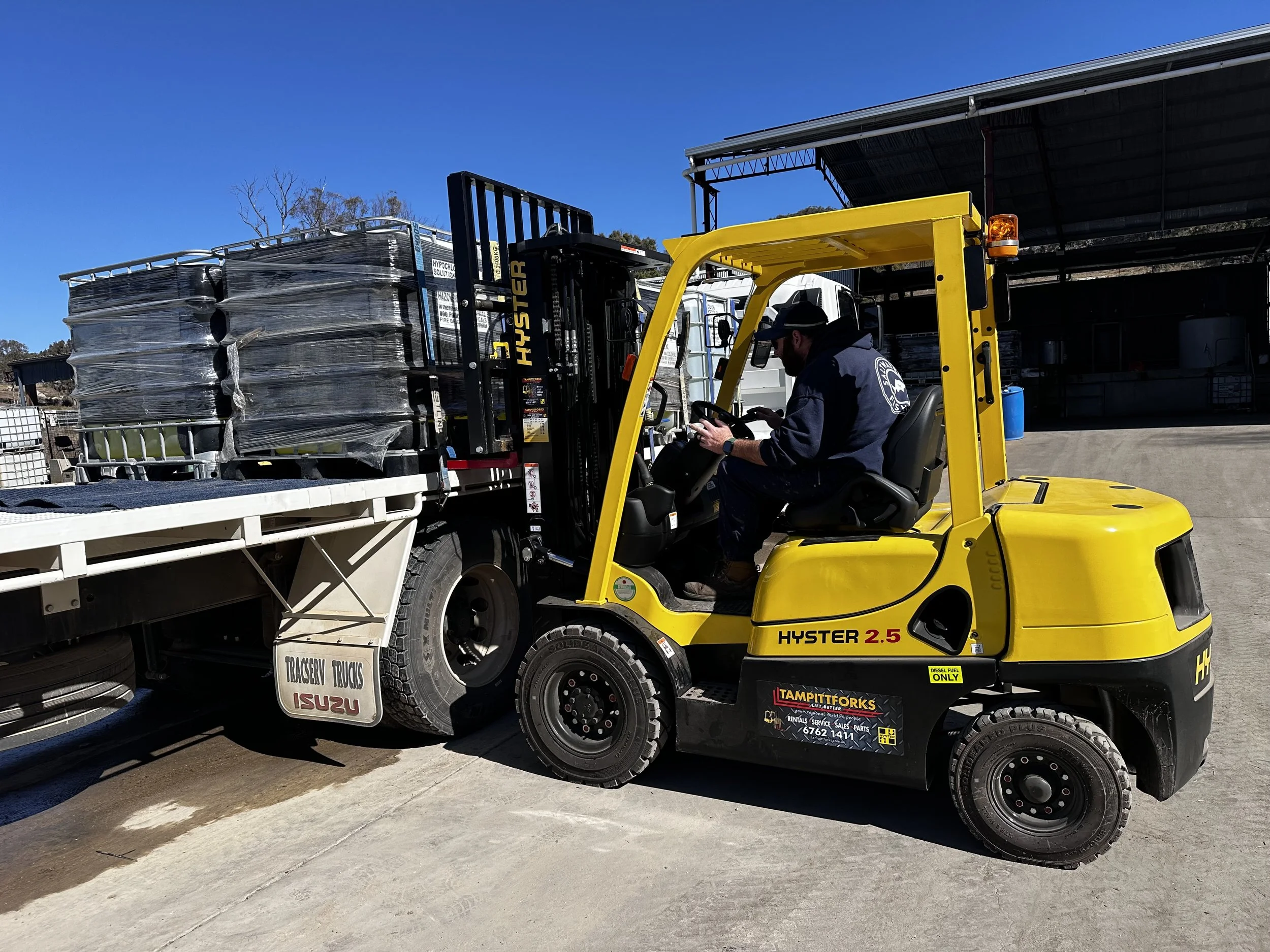 A worker operating a yellow Hyster forklift truck loads large wrapped pallets onto a flatbed truck under a clear blue sky.