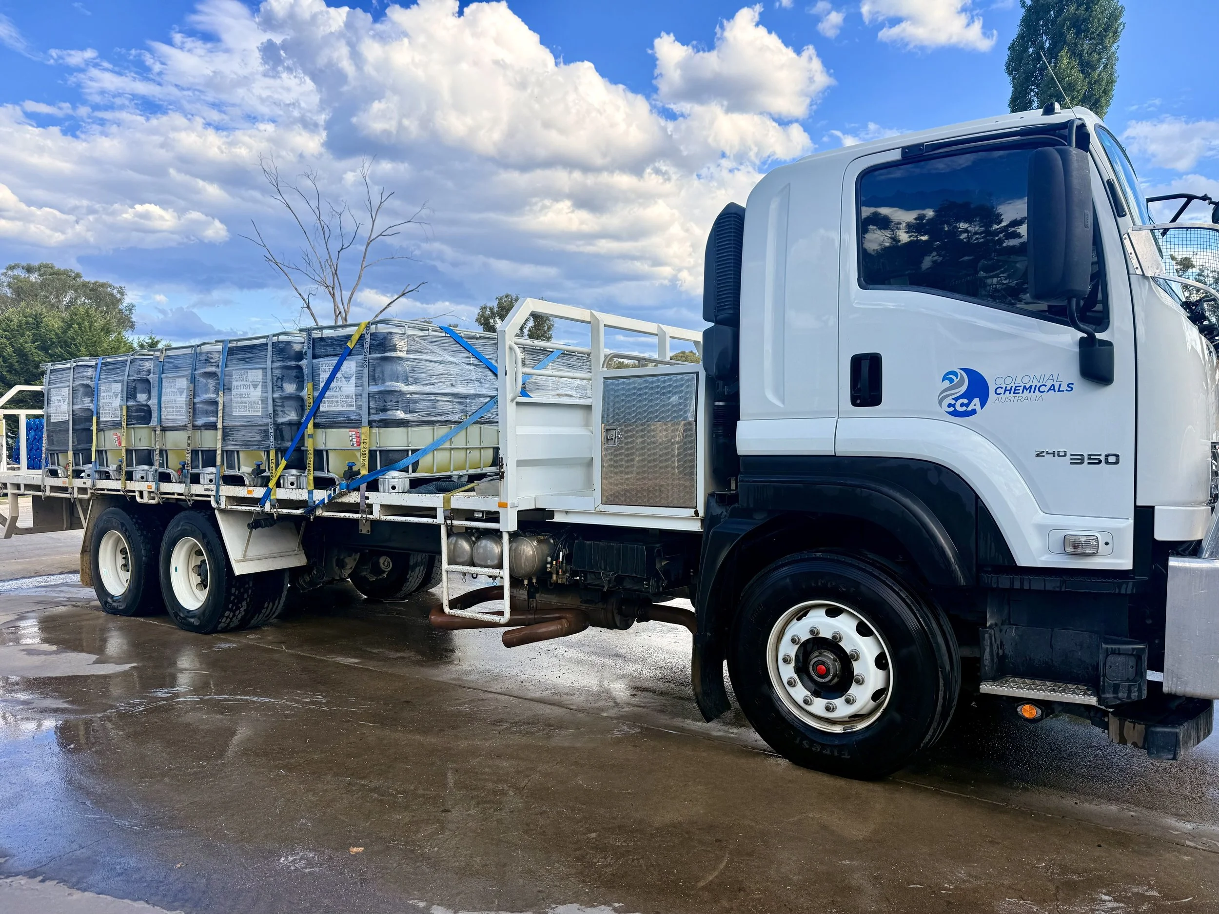 A white truck with a logo reading 'Colonial Chemicals Australia' on the door, parked outdoors under a partly cloudy sky, with some water and wet pavement around it.