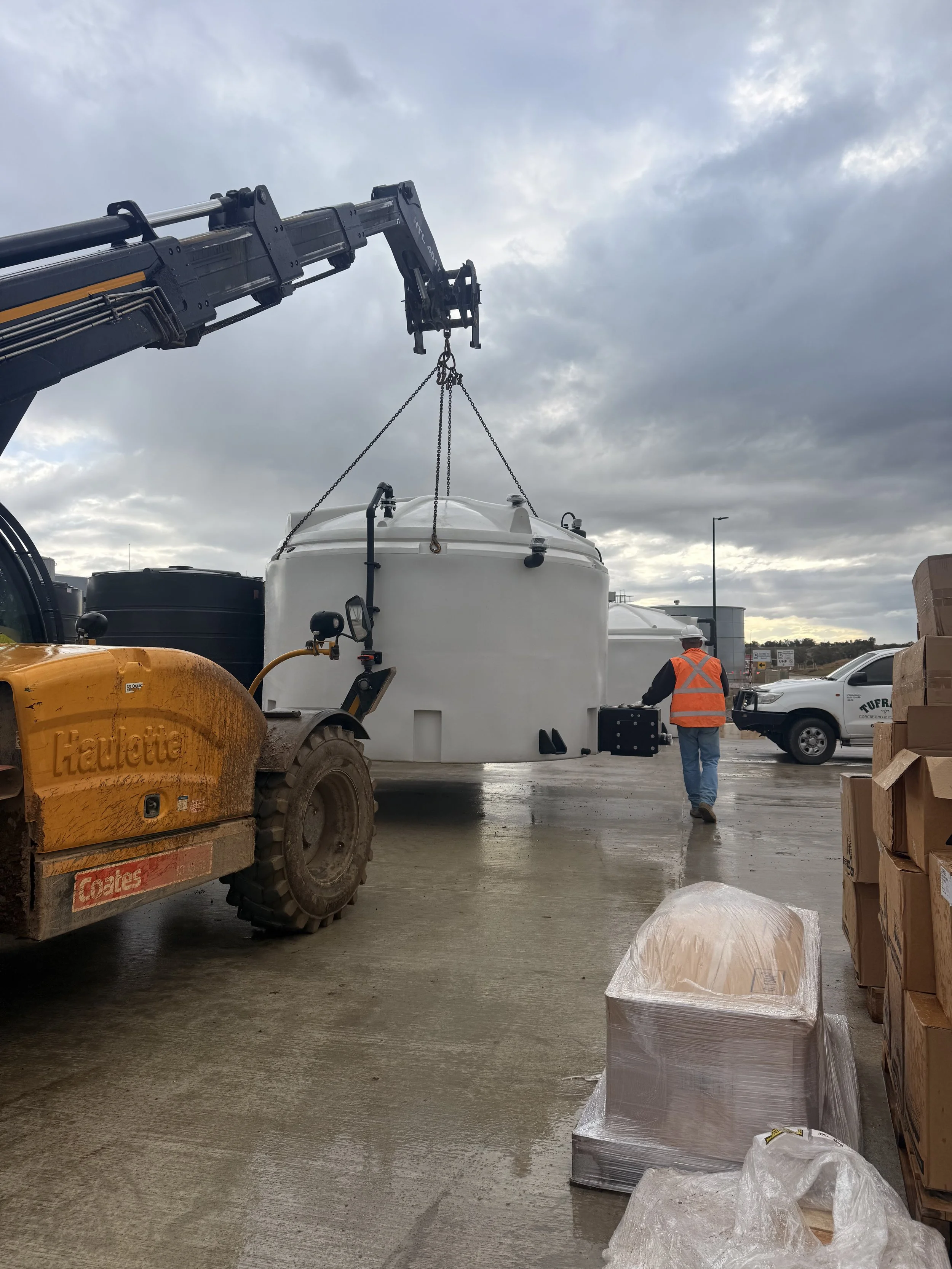 A construction site showing a lifting crane hoisting a large white tank. There are workers, a forklift, and stacked pallets with plastic wrap on the ground. Overcast sky in the background.