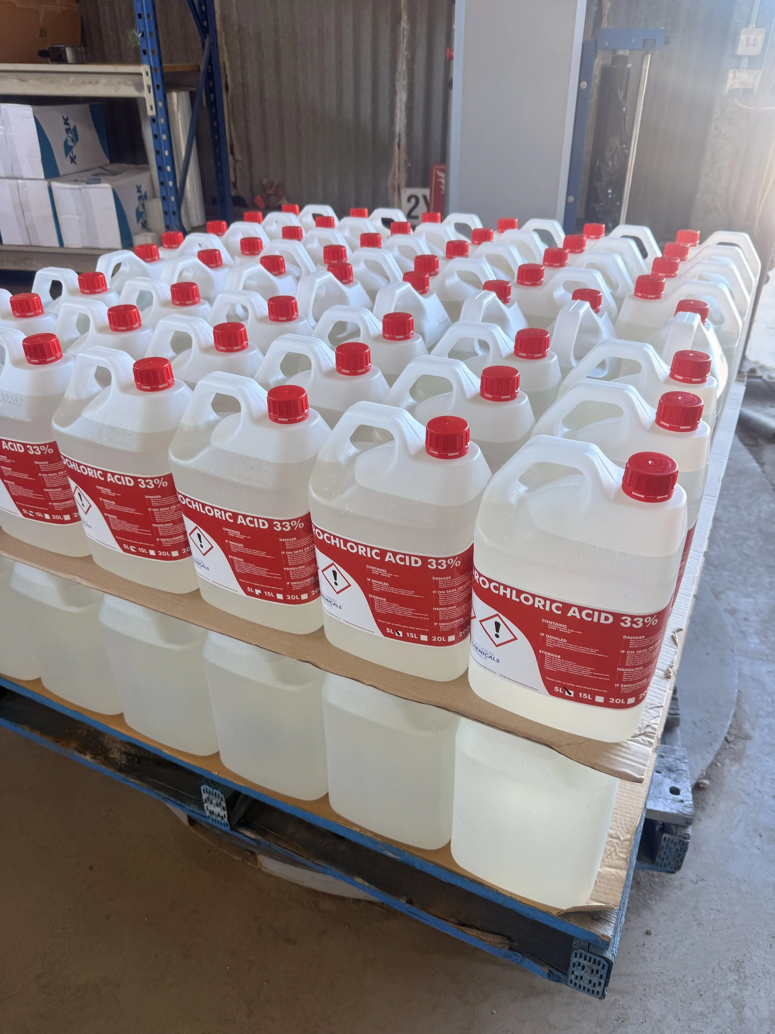 Several large bottles of 33% hydrochloric acid with red caps are arranged on a table in a workshop or storage area.