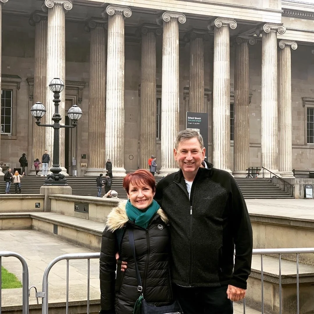A smiling man and woman standing together in front of a classical building with large columns, stairs, and an ornate street lamp, in an urban setting.