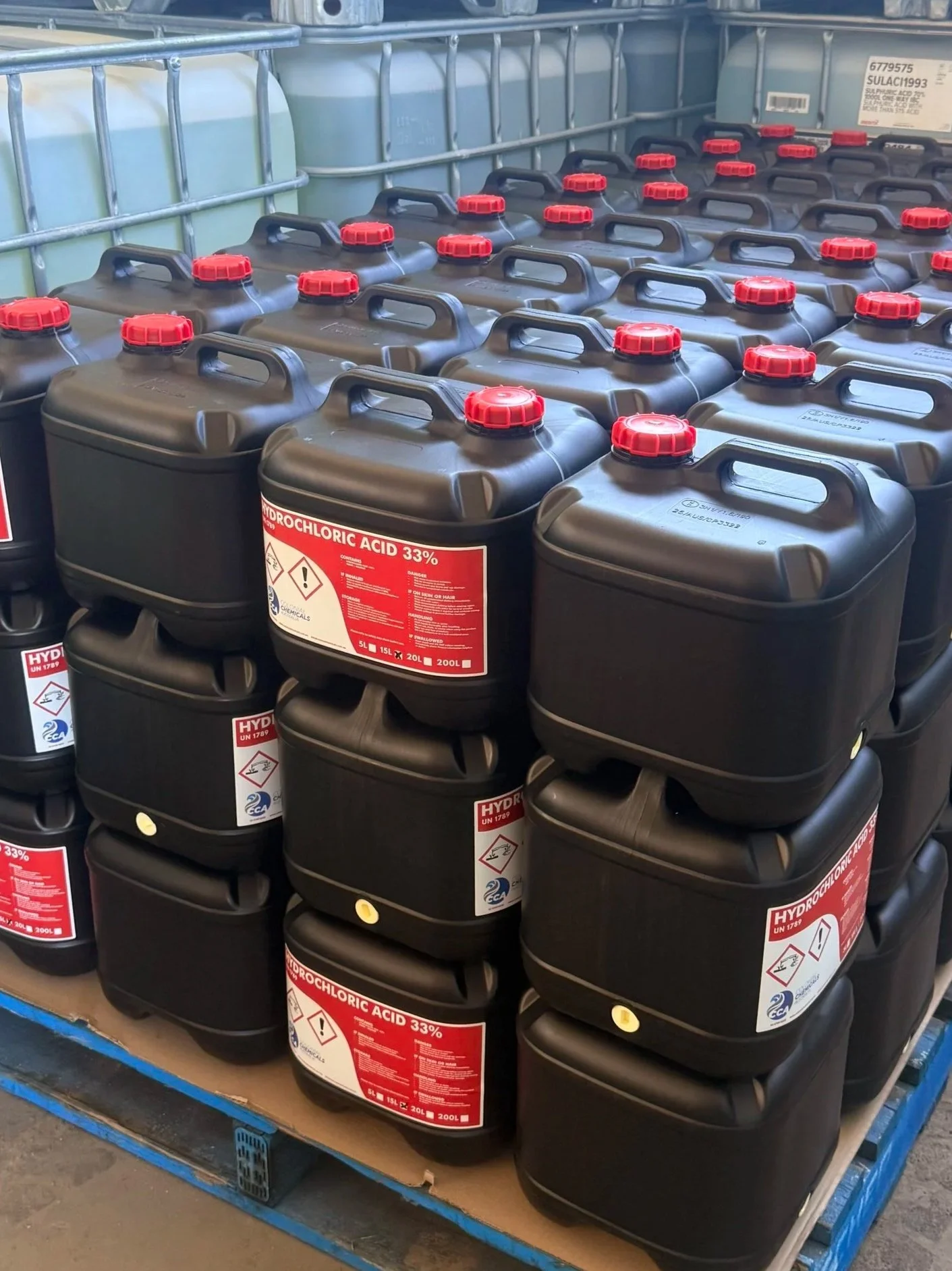 Stack of black plastic containers with red caps, labeled as containing 33% hydrochloric acid, on a blue shelving unit in a warehouse.