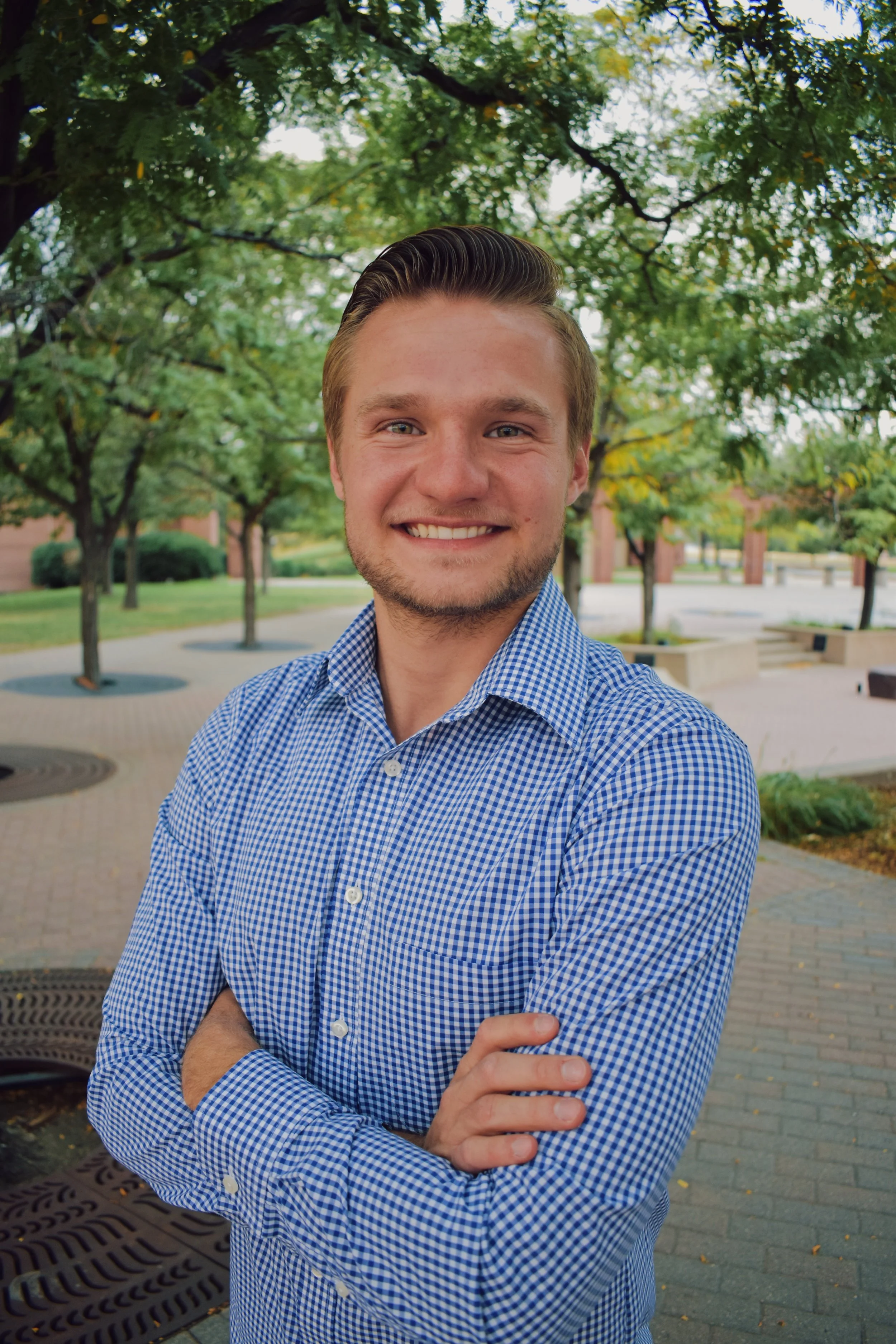 Jame(son) with short, styled brown hair and a beard, wearing a blue and white checkered shirt, smiling with his arms crossed in an outdoor park setting with green trees and brick paths.