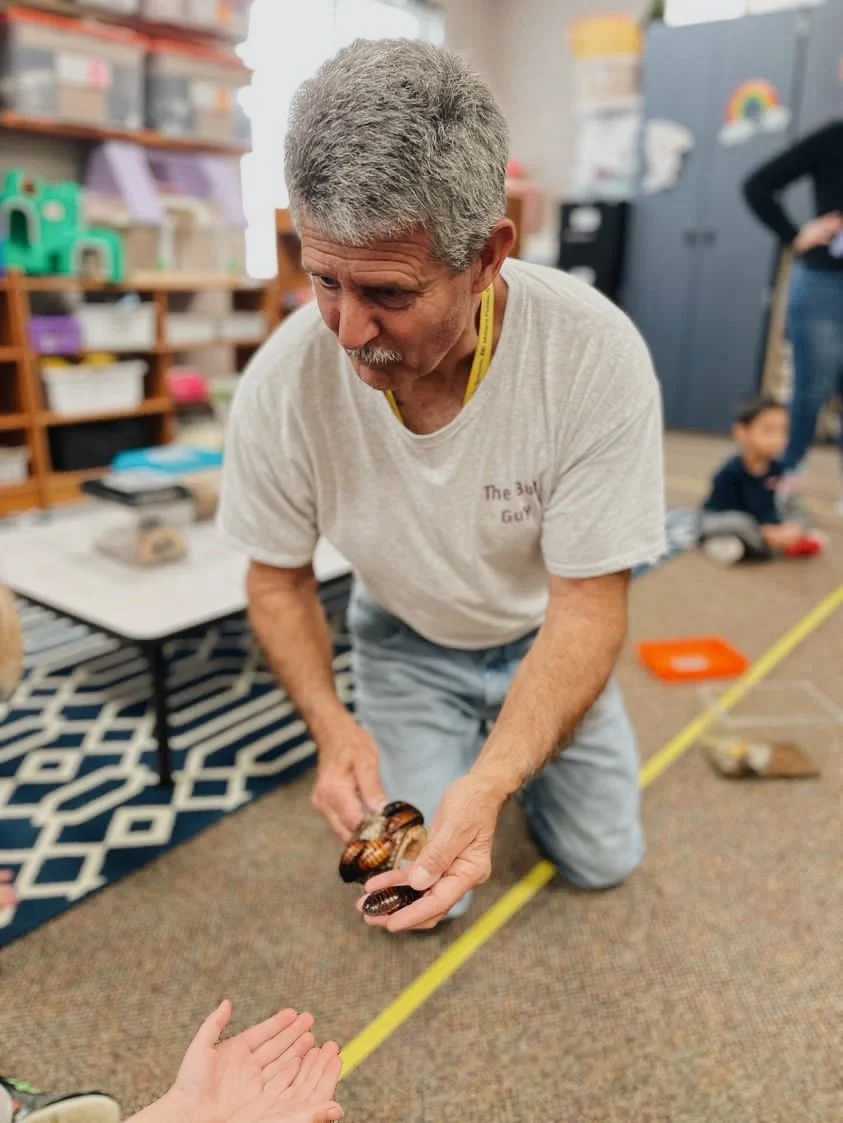 The Bug Guy kneeling on the floor of a classroom, holding a hissing cockroach with both hands as he shows it to a child.