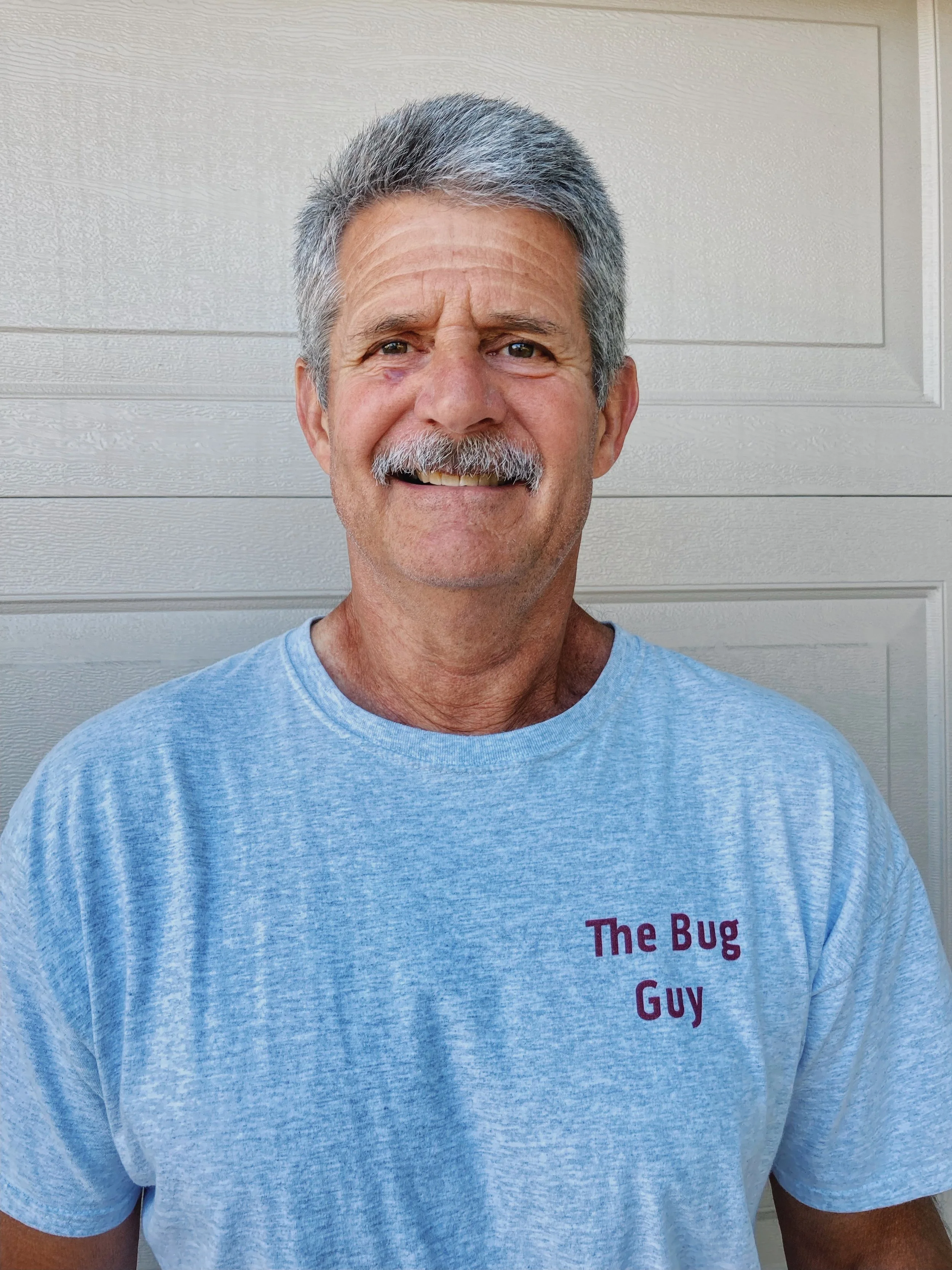 The Bug Guy, smiling, wearing a light gray t-shirt with text that partially reads "The Bug Guy," standing in front of a beige wall.